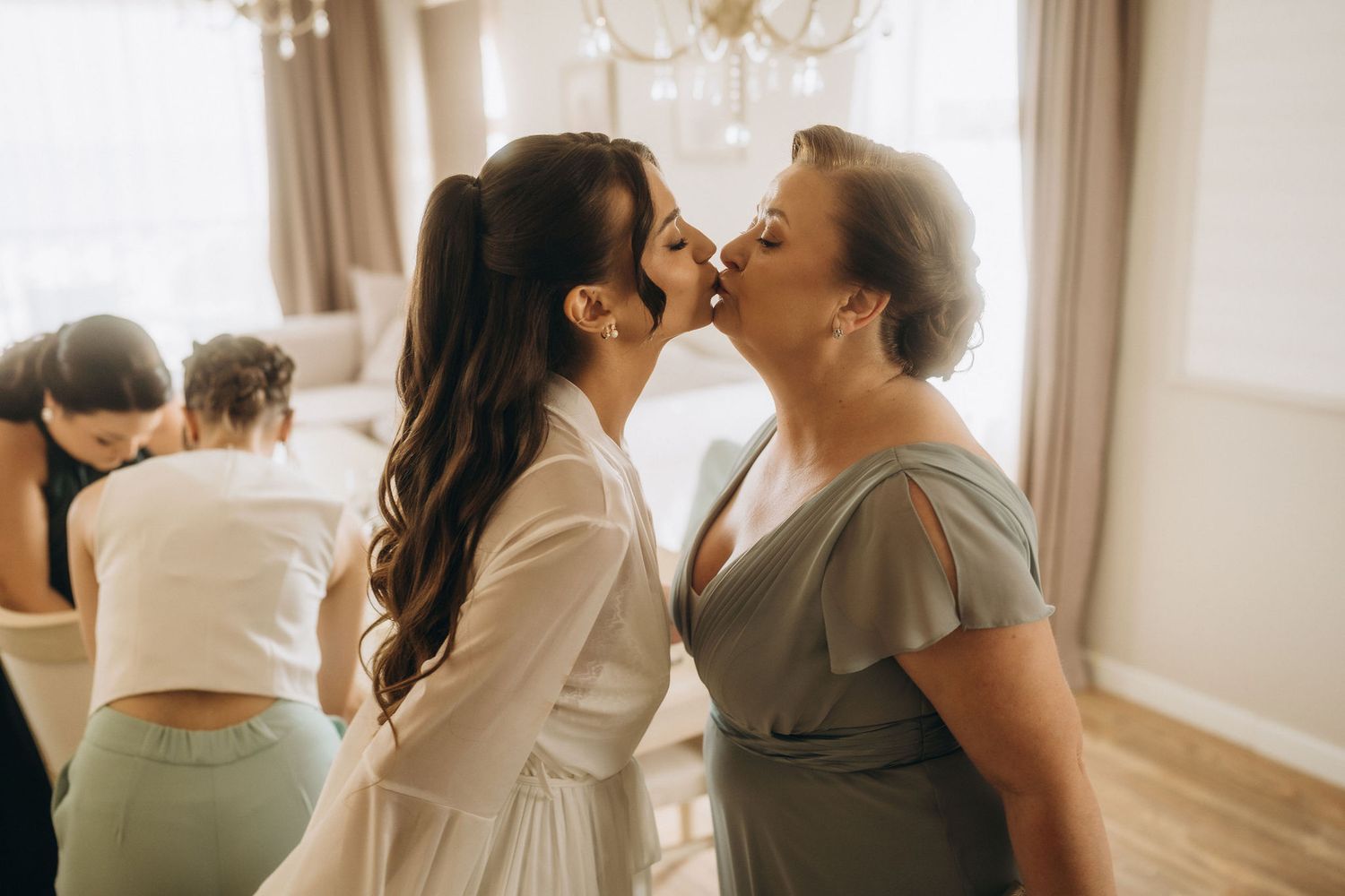 Bride and mother sharing a tender kiss during wedding preparations in a bright elegant room with bridesmaids in background.