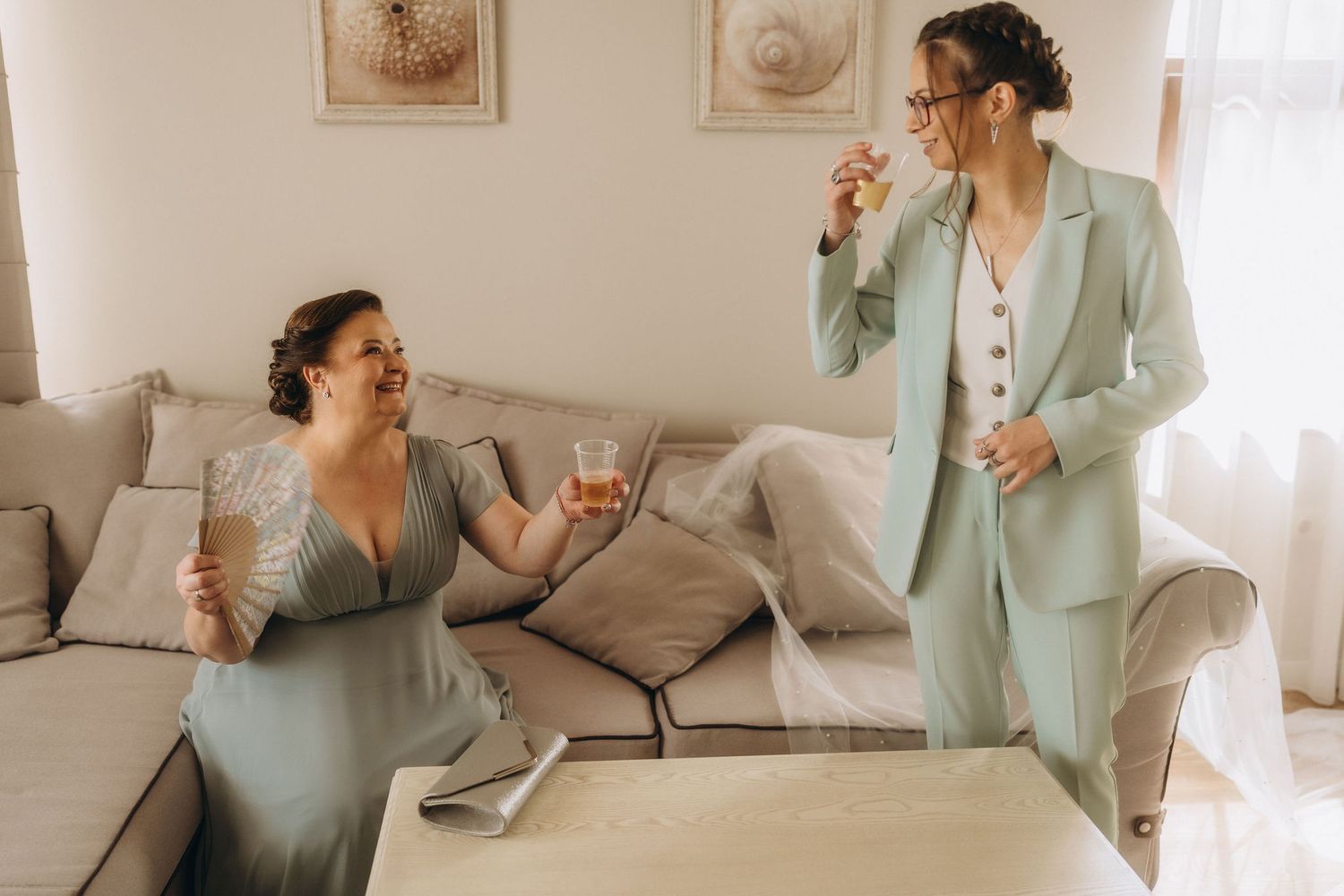 Two women in elegant attire toast with drinks in a bright living room during a wedding celebration.