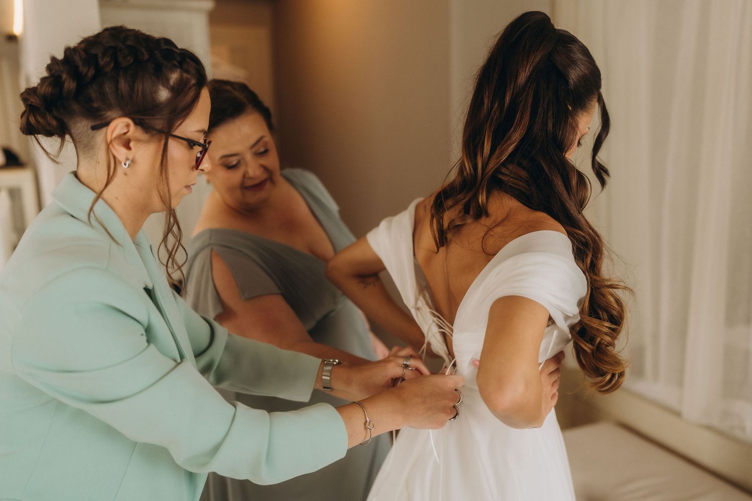 Two bridesmaids in sage green help a bride in a white off-shoulder gown with her dress buttons before her wedding ceremony.