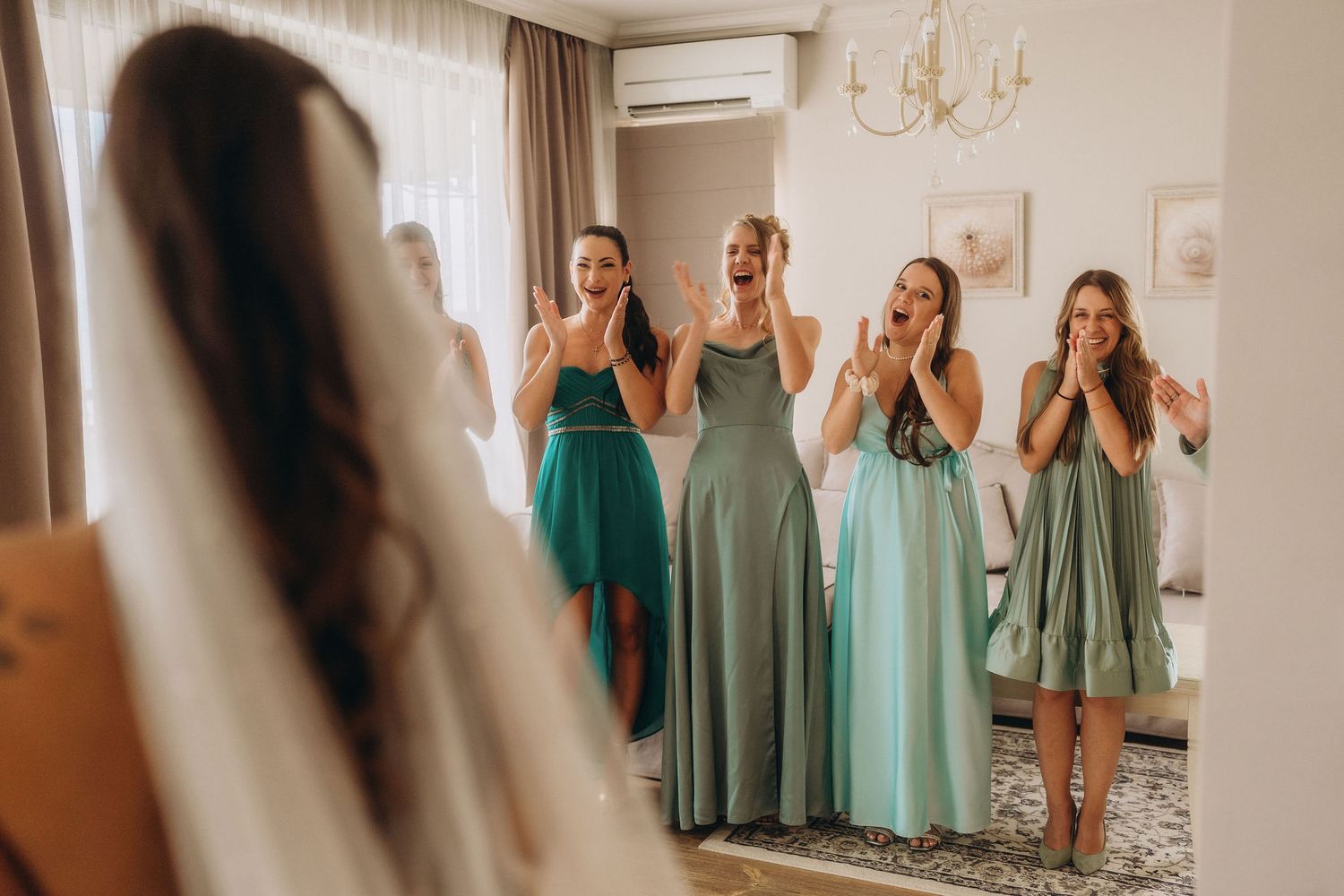 Bridesmaids in green dresses react with joy as bride reveals her wedding gown in a bright, elegant room.