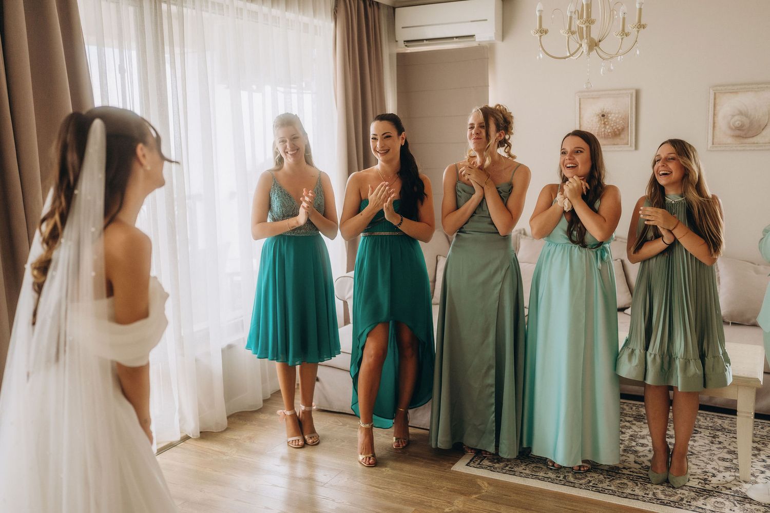 Bride in white gown reveals look to five bridesmaids in green dresses, who react with joy and excitement indoors.