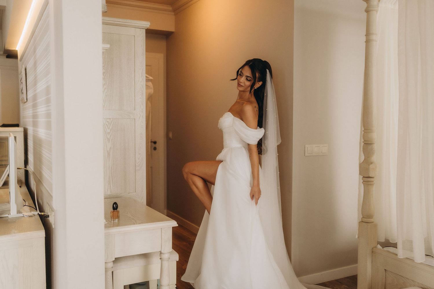 Elegant bride in off-shoulder white wedding gown with veil poses gracefully in a softly lit bridal suite.