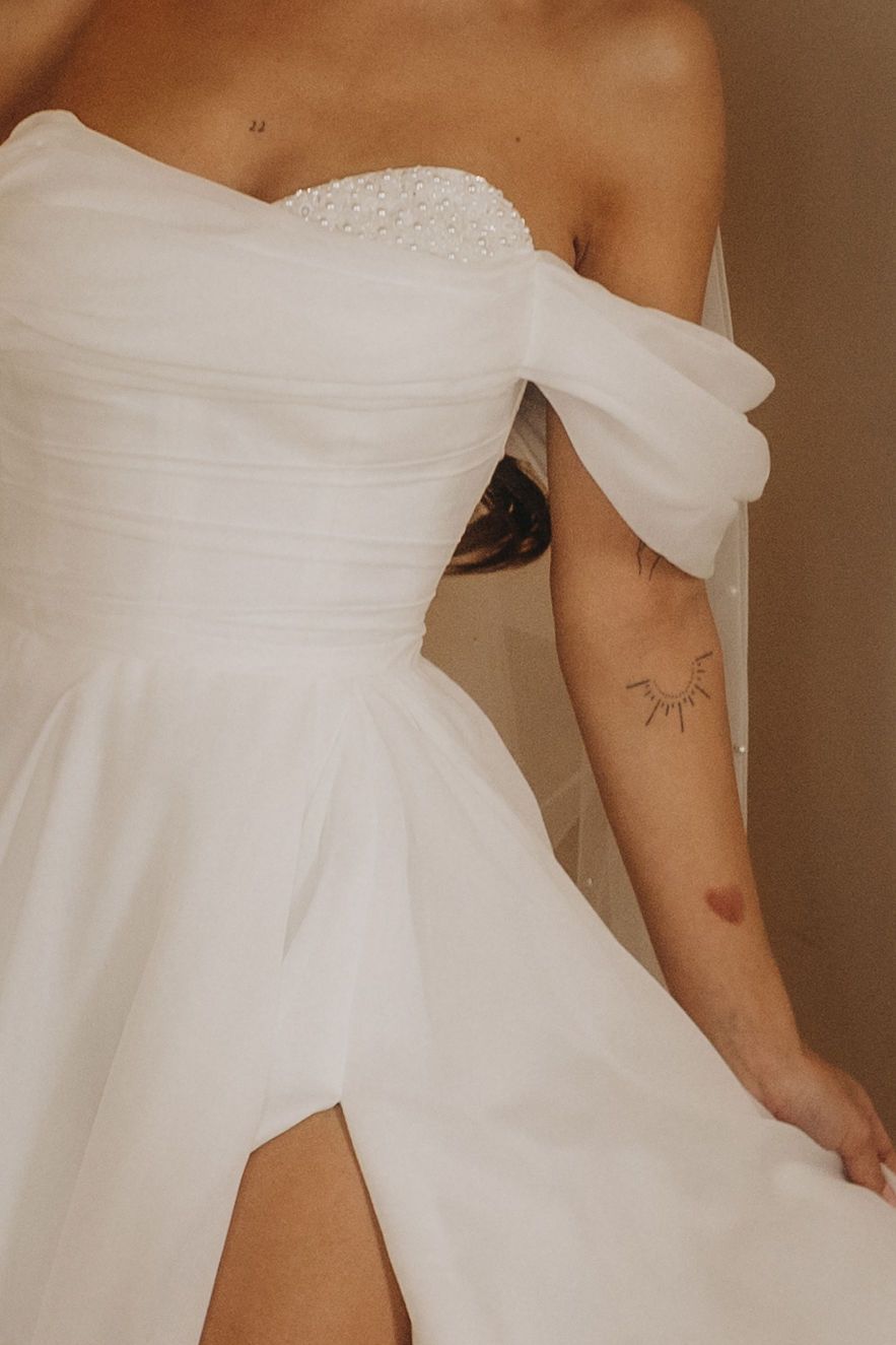 Elegant off-shoulder white wedding dress with ruched bodice, beaded detail, and thigh-high slit on a bride.