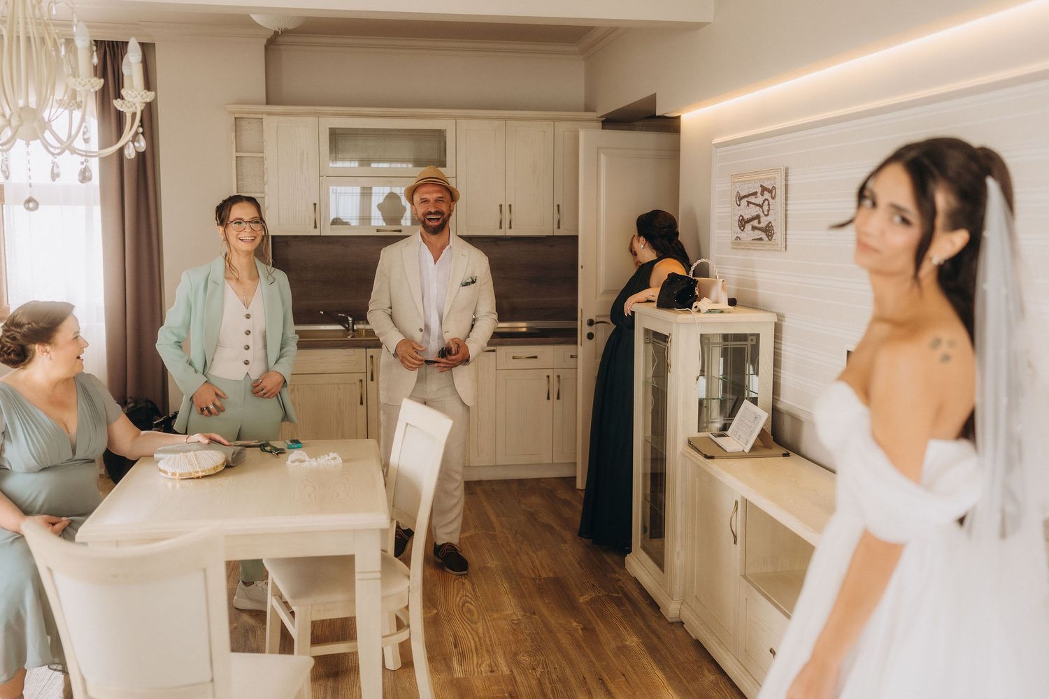 Bride in white gown stands in elegant kitchen while family members gather around a dining table during wedding preparations.