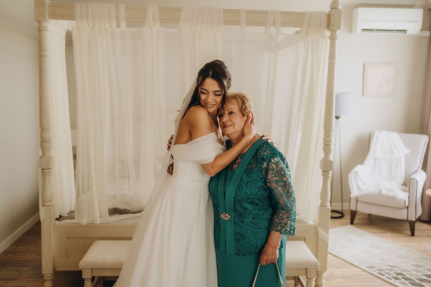 Bride in white off-shoulder gown hugging older woman in teal lace dress inside elegant white bedroom.