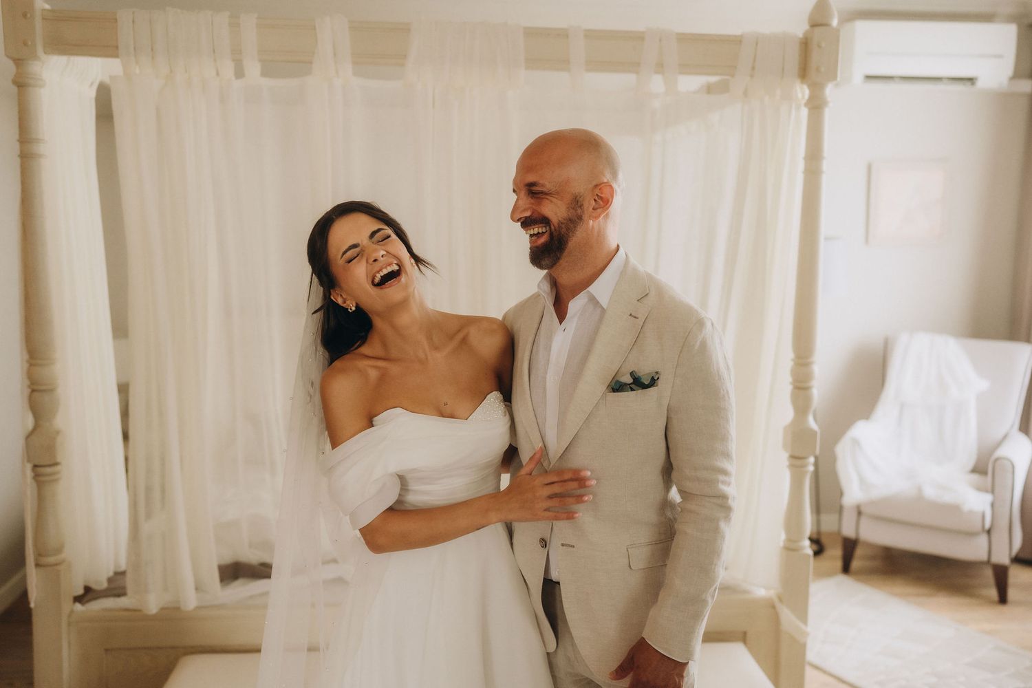 Joyful bride and groom laughing together in elegant white room on wedding day, bride in strapless gown, groom in tan suit.