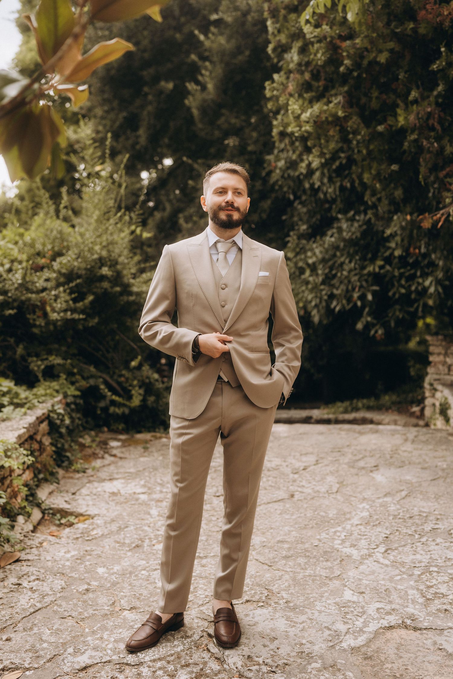 Groom in beige suit with tie stands confidently in lush garden setting for outdoor wedding portrait.