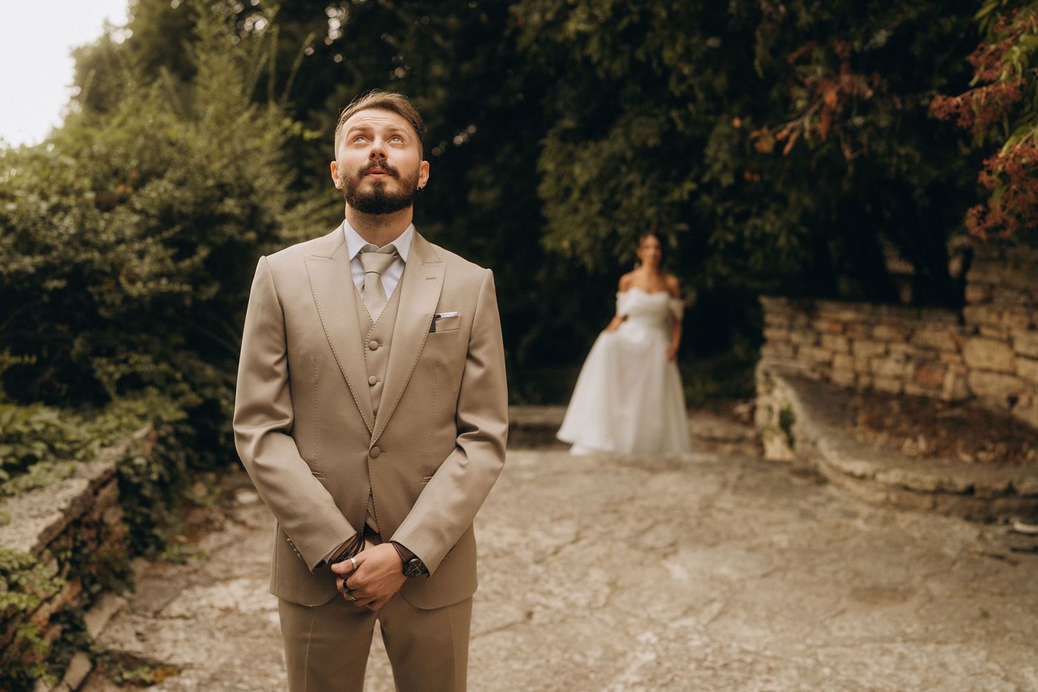 Groom in beige suit stands in garden path as bride in white wedding dress poses softly blurred in background.