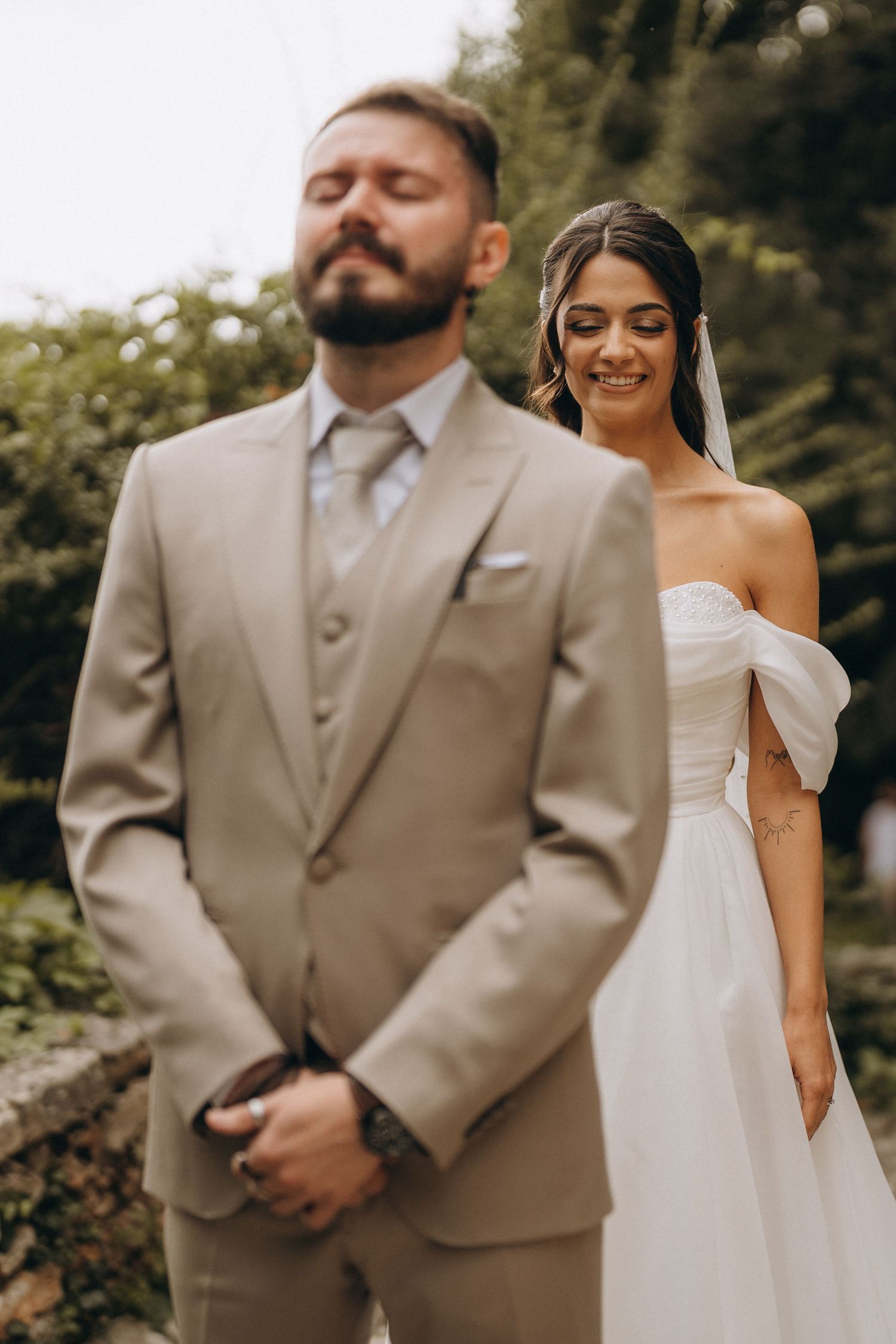 A smiling bride in a white off-shoulder gown stands behind her bearded groom in a tan suit during an outdoor wedding.