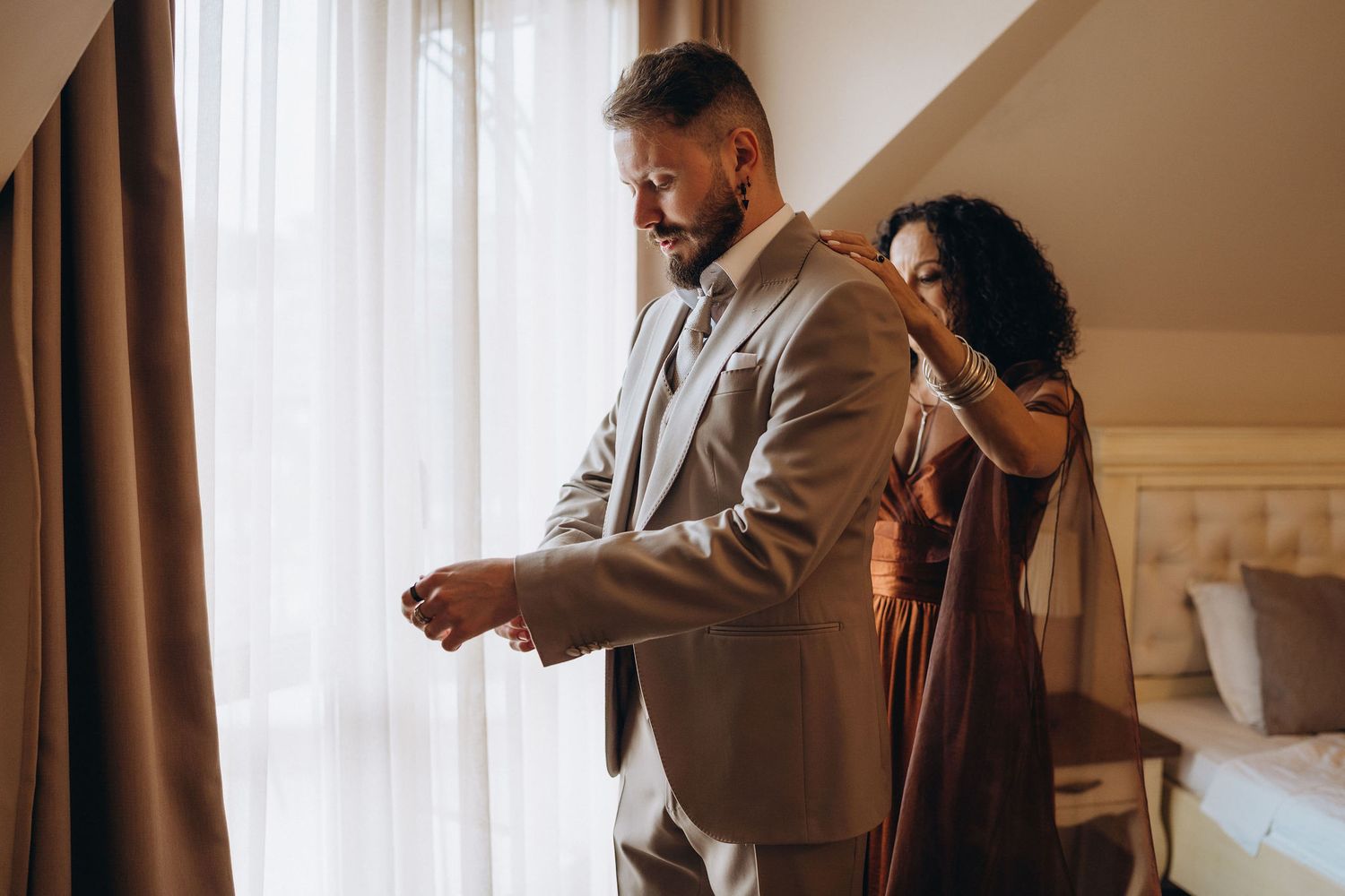 A woman in a brown dress helps a bearded man in a gray suit adjust his cufflinks in an elegant hotel room.