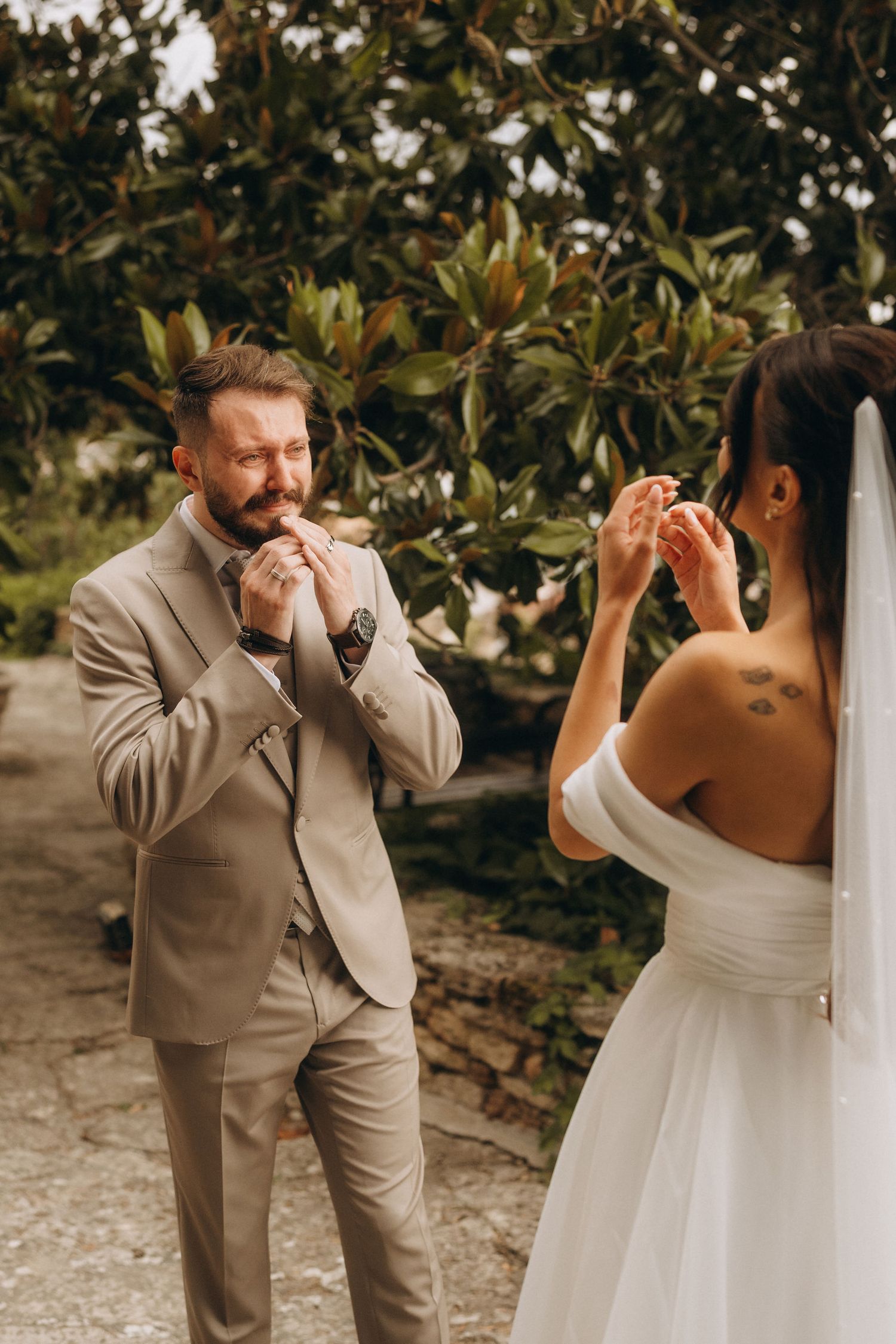 Emotional groom in beige suit reacts to seeing bride in white off-shoulder wedding dress outdoors near lush green trees.
