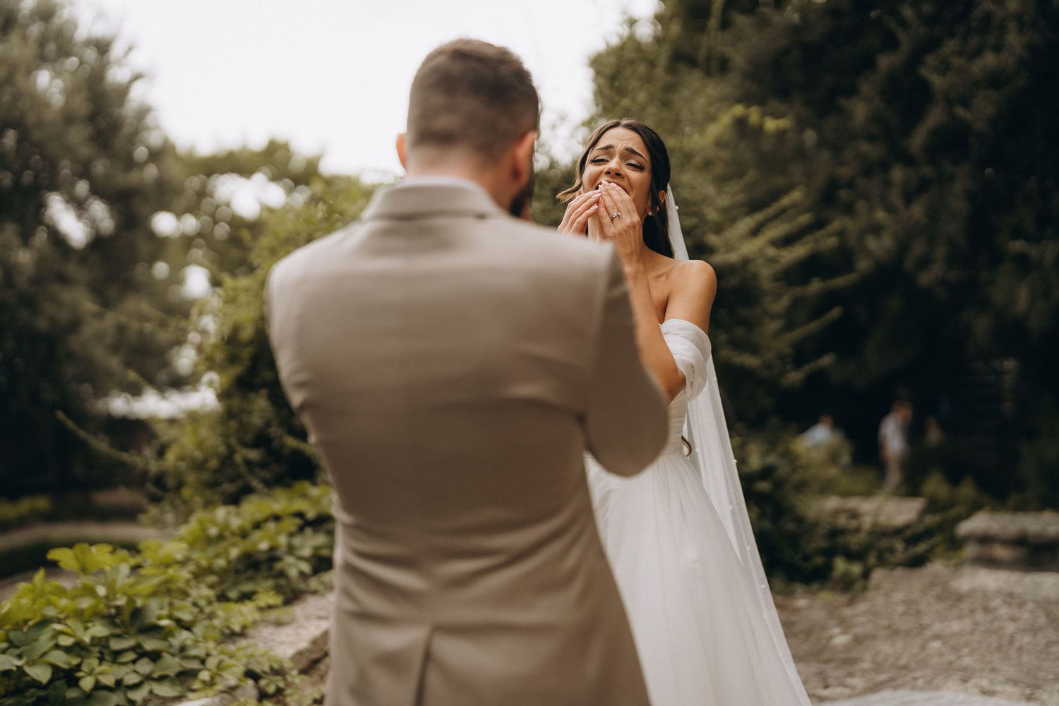 Emotional bride in white gown covers mouth during first look with groom in tan suit in lush garden setting.