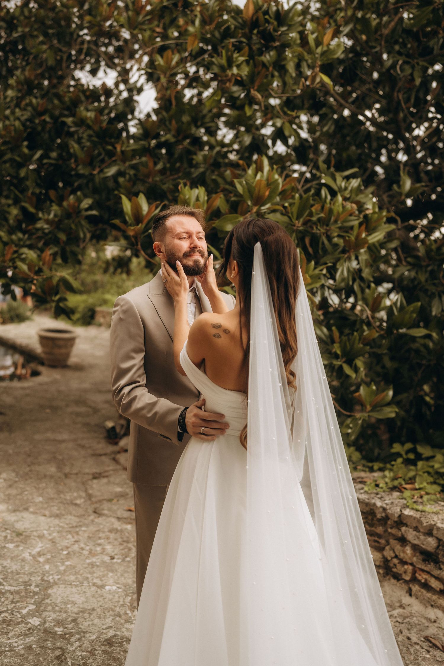 Bride in white gown and veil tenderly holds groom's face in lush garden setting during romantic outdoor wedding.