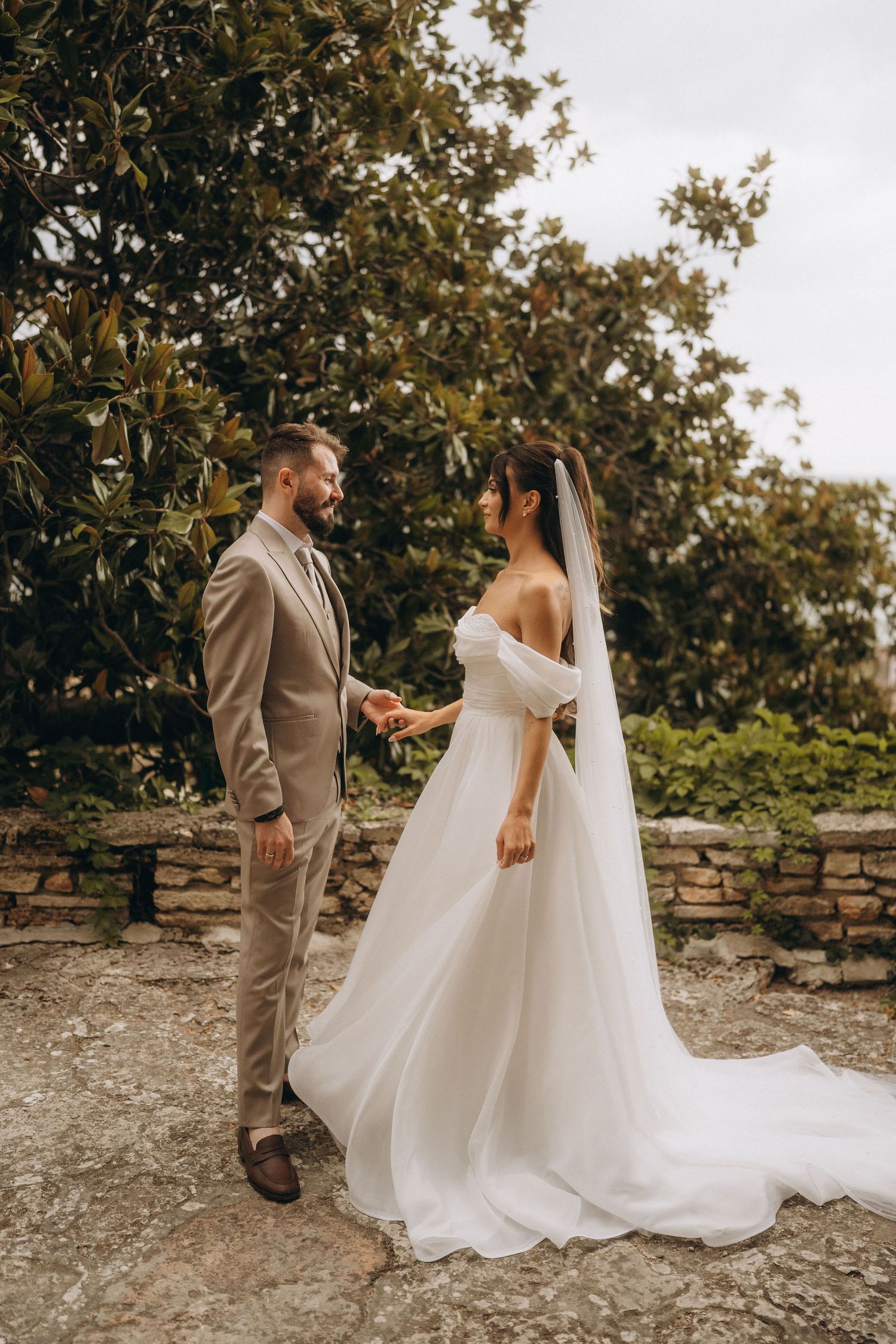 Bride in white off-shoulder gown with veil holds hands with groom in beige suit outdoors.