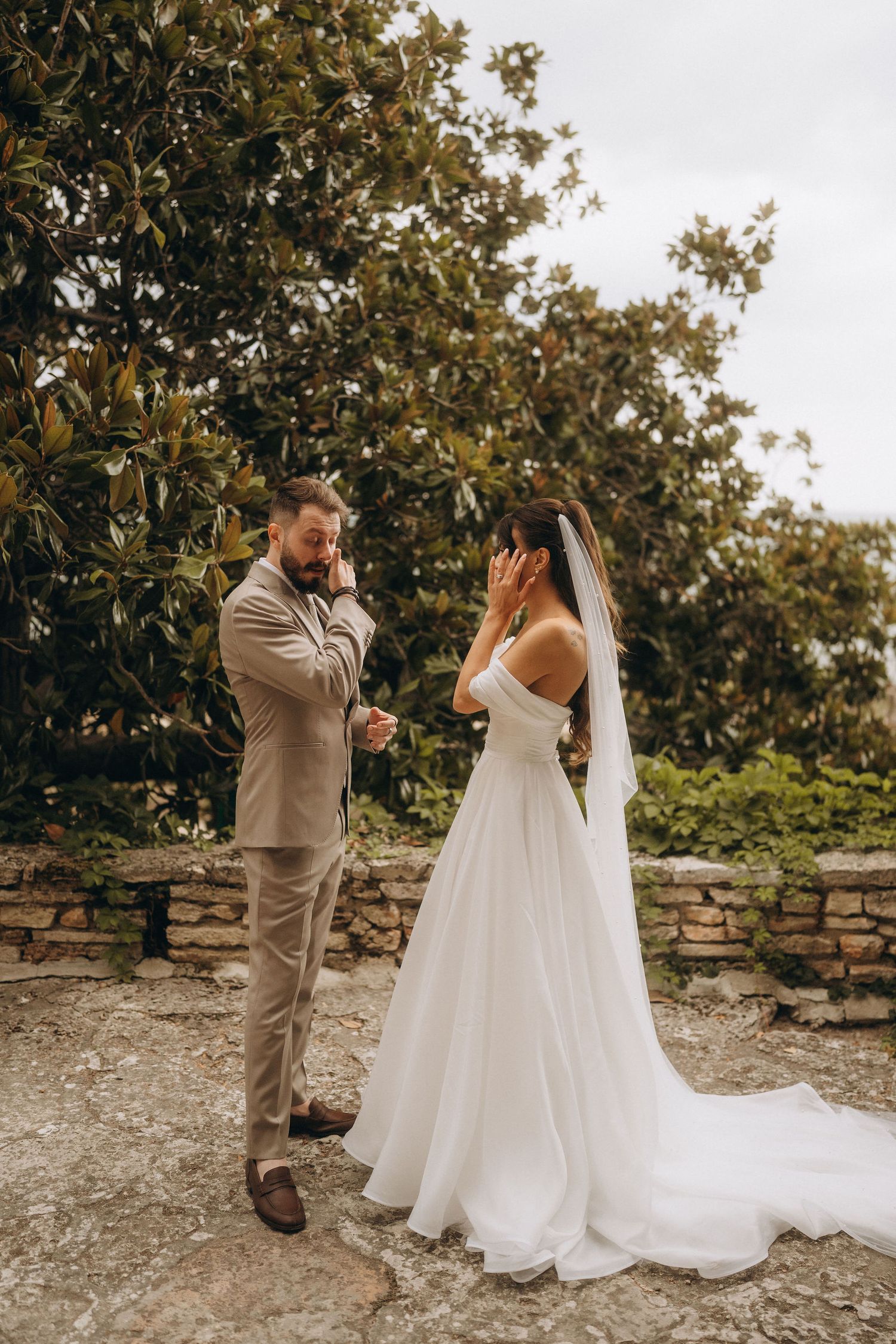 Emotional bride and groom wiping tears during outdoor wedding ceremony near stone wall.
