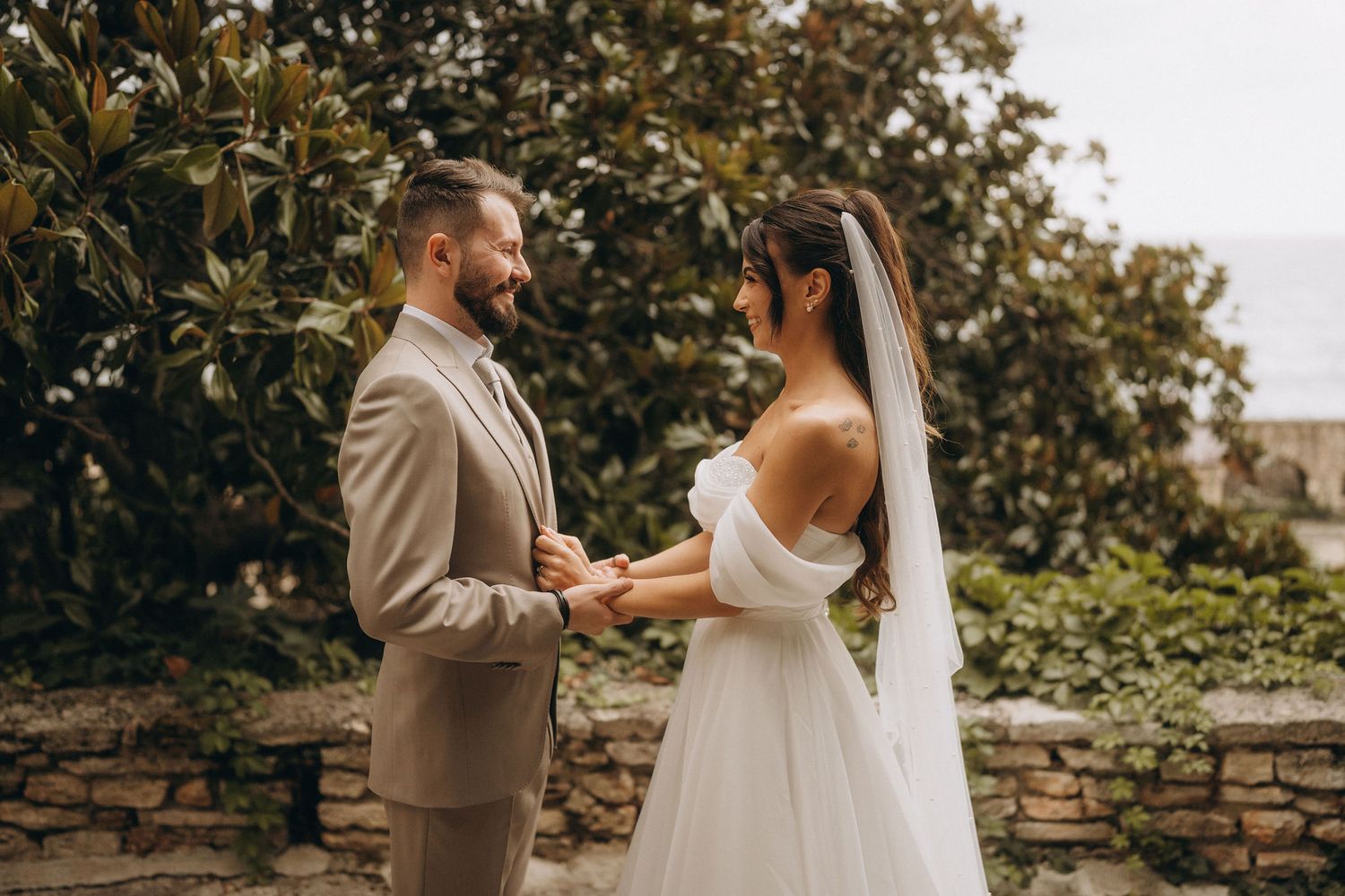 A bride and groom hold hands outdoors in a garden setting, with the groom in a tan suit and bride in a white off-shoulder gown.
