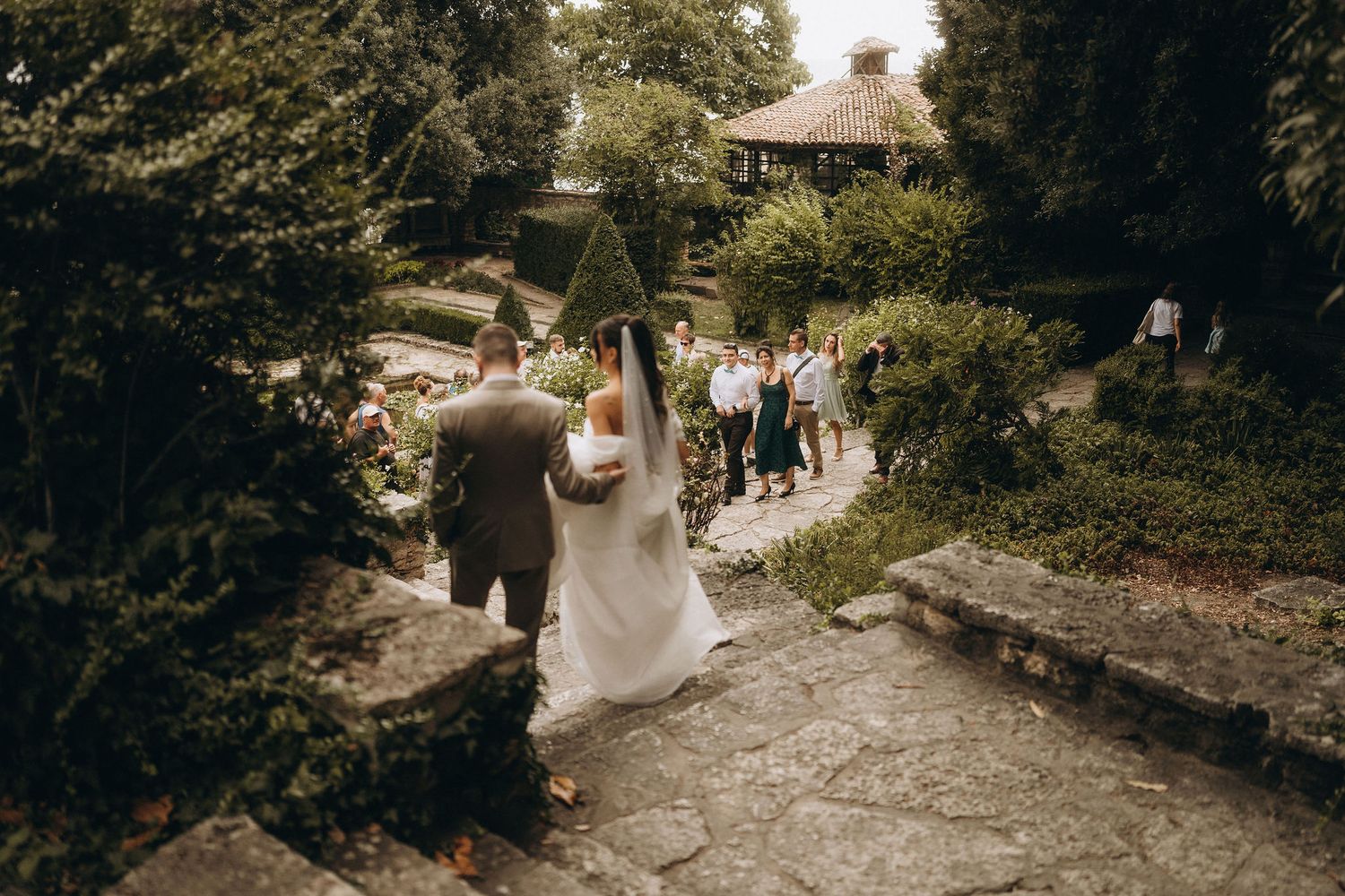 Bride and groom walking down stone steps in a lush garden wedding venue with guests gathered near a gazebo.