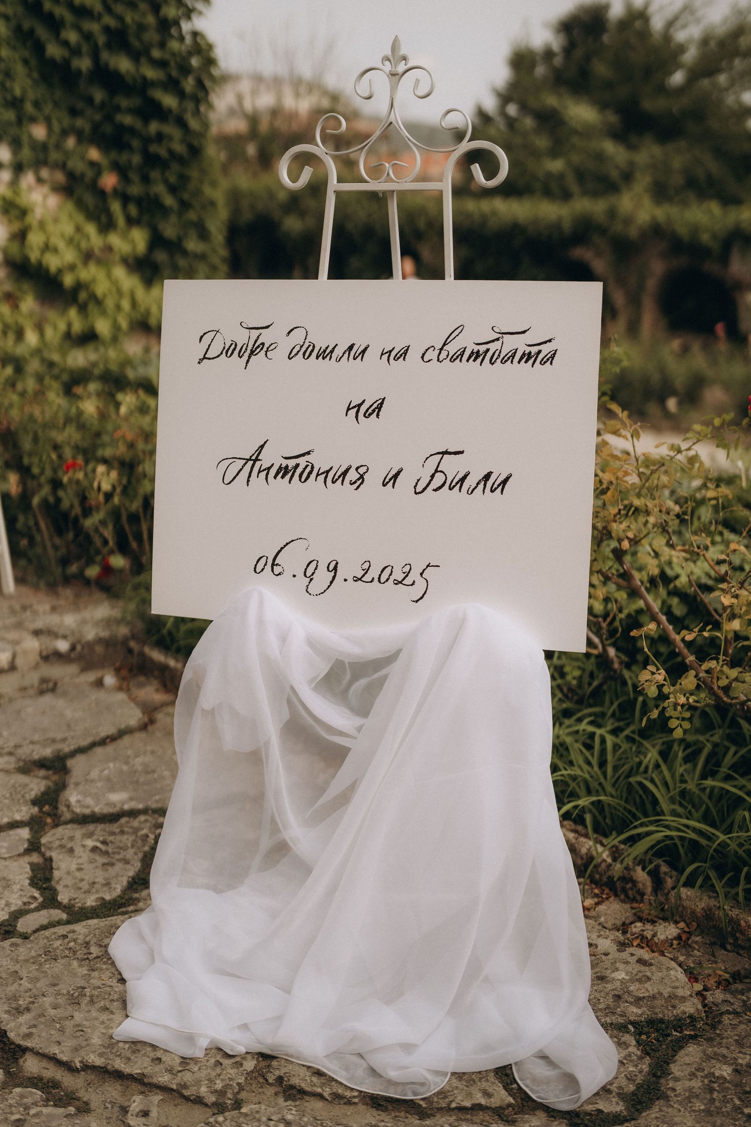Elegant wedding welcome sign in Bulgarian on a white easel with flowing tulle, dated 06.09.2025, set in a garden.