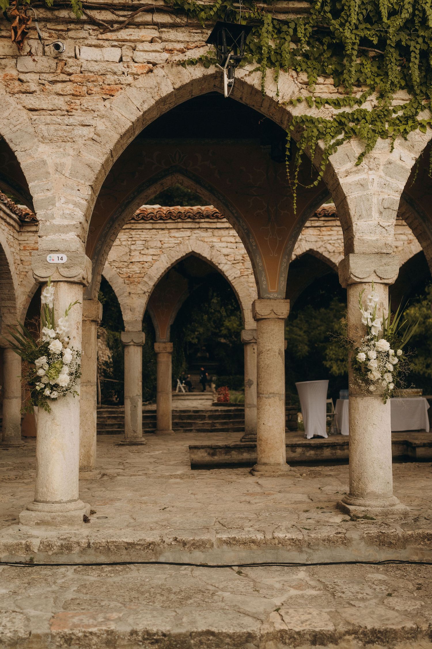 Stone archway colonnade with floral arrangements decorating columns at an elegant outdoor wedding venue.