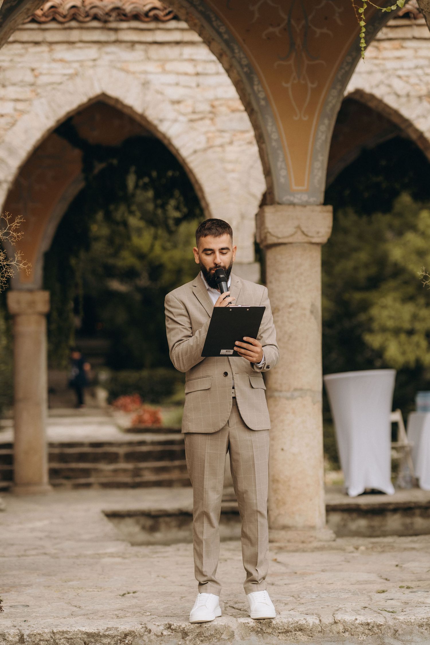 Bearded man in beige suit reads from clipboard outdoors near stone arches at a wedding venue.