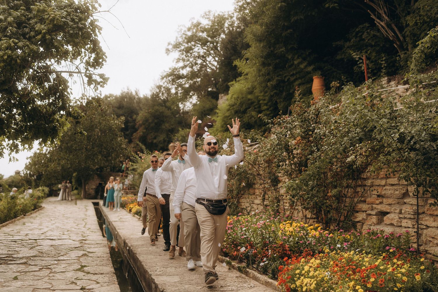 Groomsmen in white shirts and khaki pants walk along a garden path, with one man waving and smiling at a wedding.