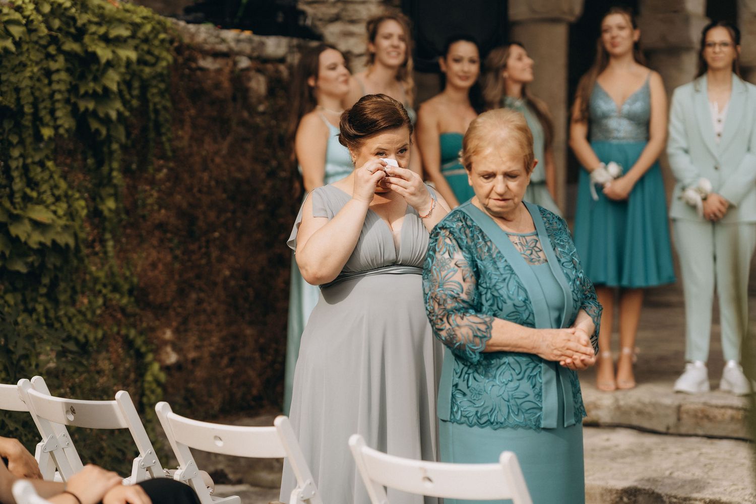 Emotional wedding guests in blue dresses crying during outdoor ceremony with bridesmaids standing in background.