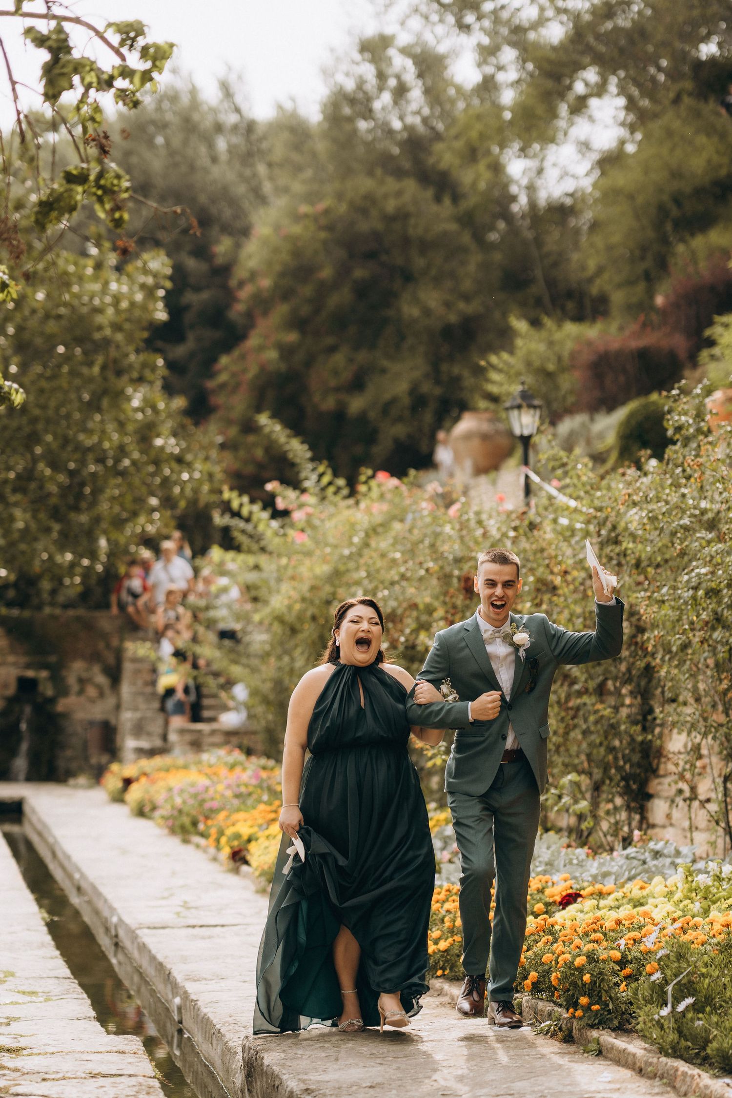 A joyful bridesmaid in black gown and groomsman in gray suit walk through a lush garden path at a wedding.