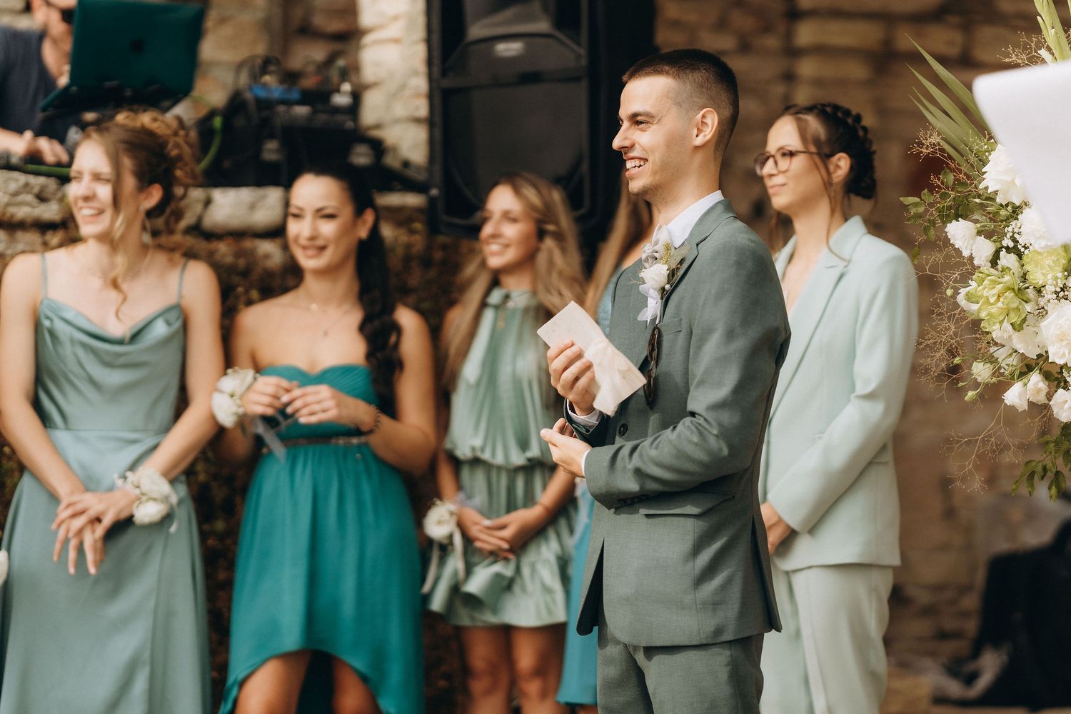 Groom in gray suit smiling at wedding ceremony with bridesmaids in teal dresses standing in background.