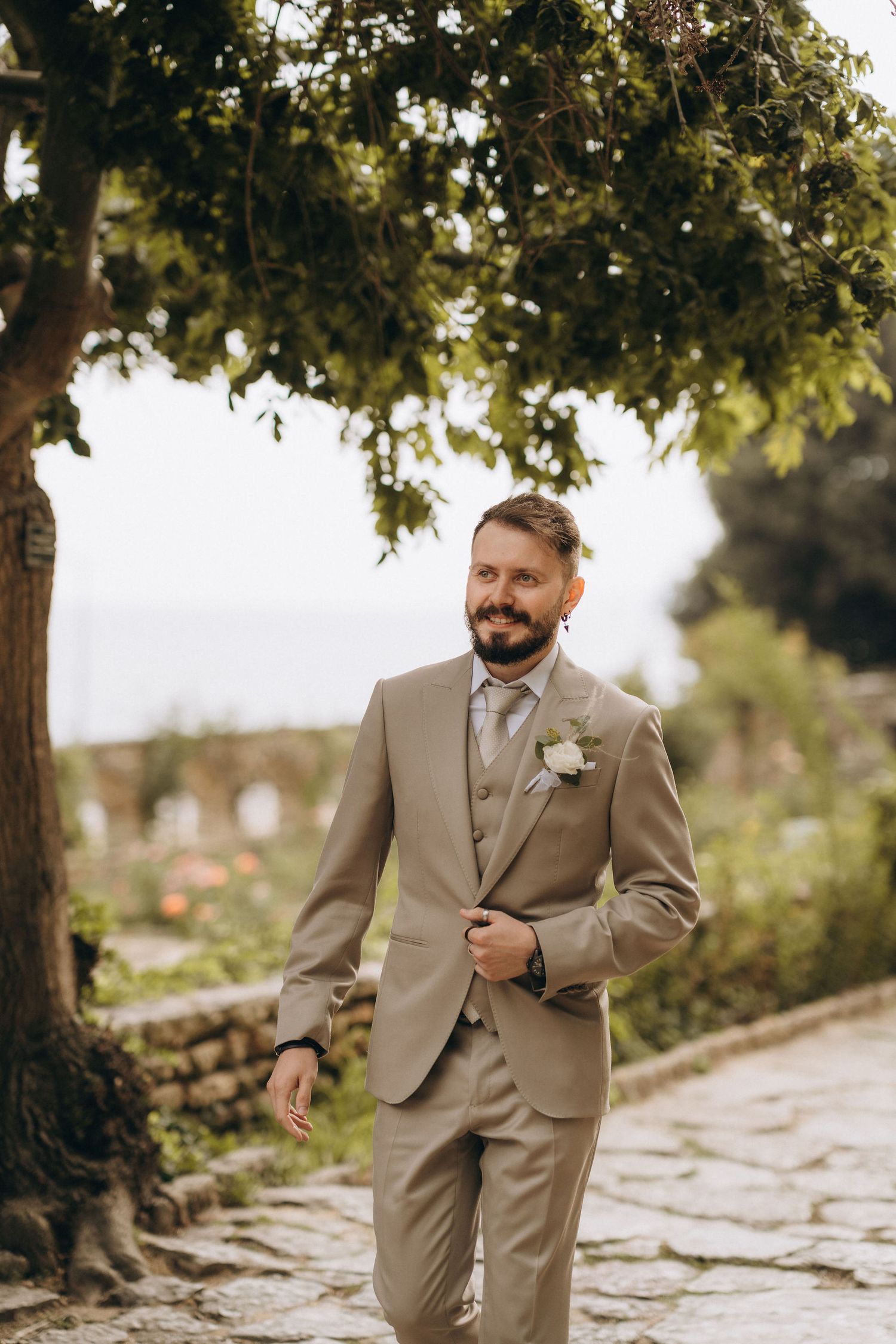 A bearded groom in a tan three-piece suit with boutonniere walks along a stone path under a lush green tree.