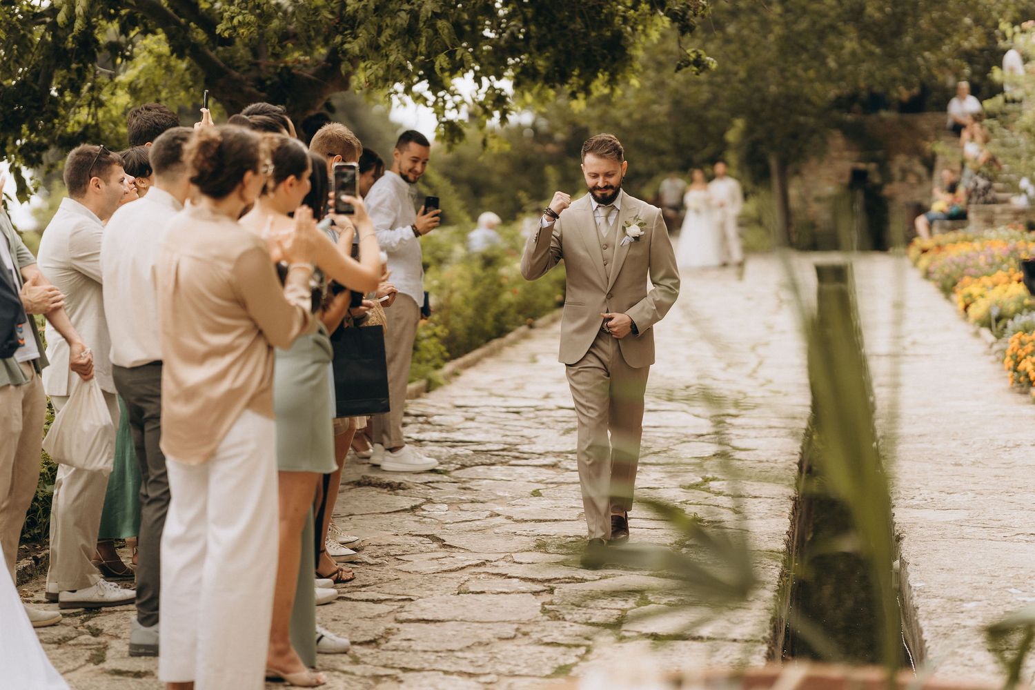 Groom in tan suit walks down outdoor stone aisle as wedding guests applaud in a lush garden ceremony.