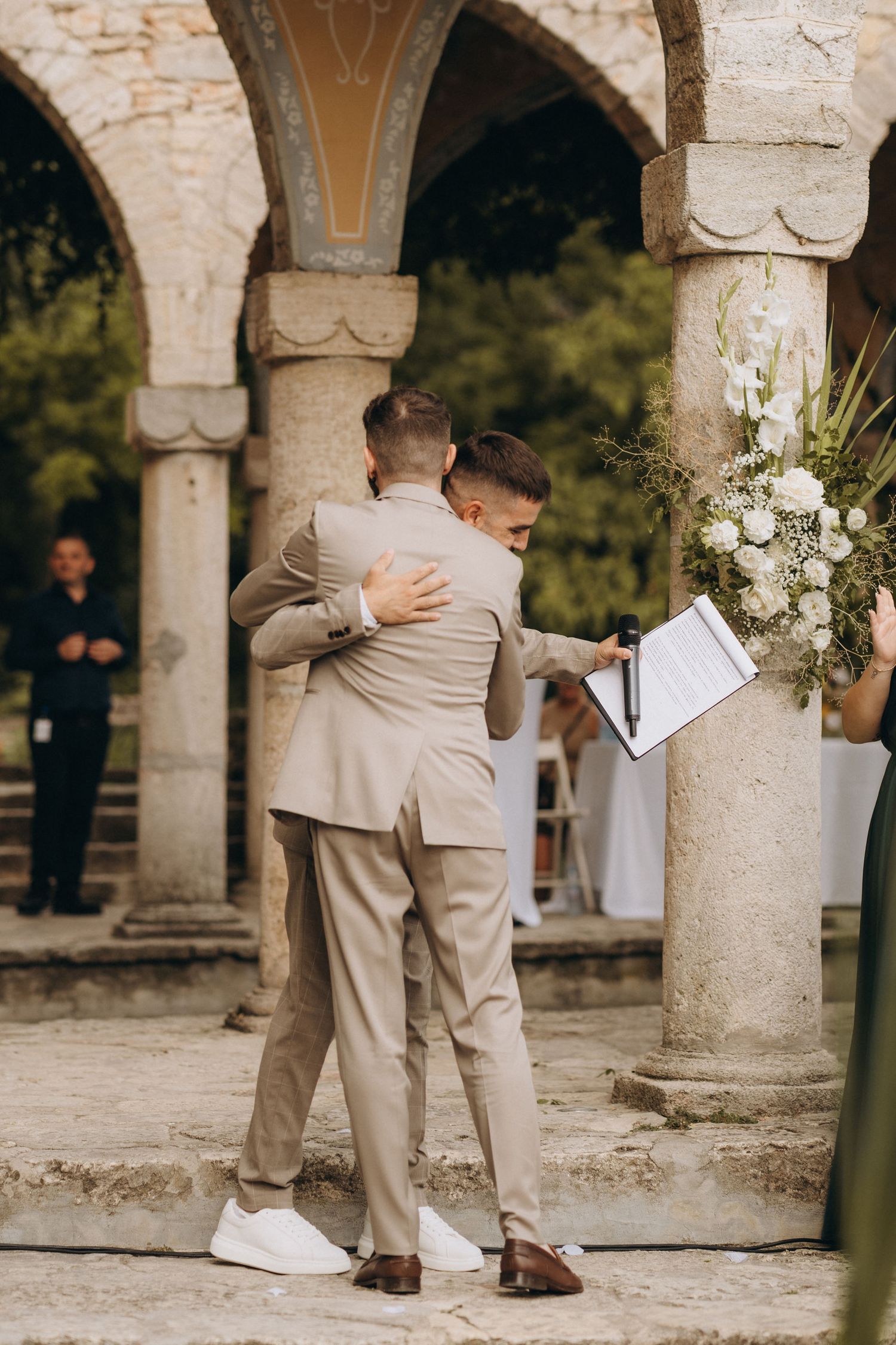 Two grooms in matching beige suits share an emotional hug during their outdoor wedding ceremony among stone columns.