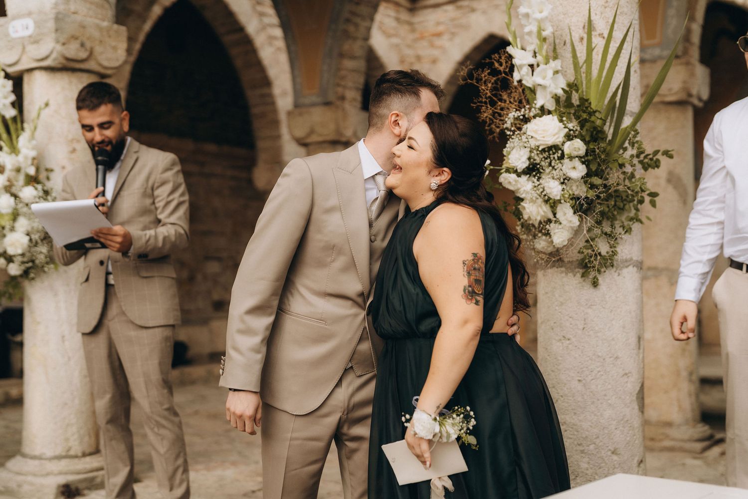 Couple sharing a romantic moment at an outdoor wedding ceremony with floral arrangements and stone architecture.