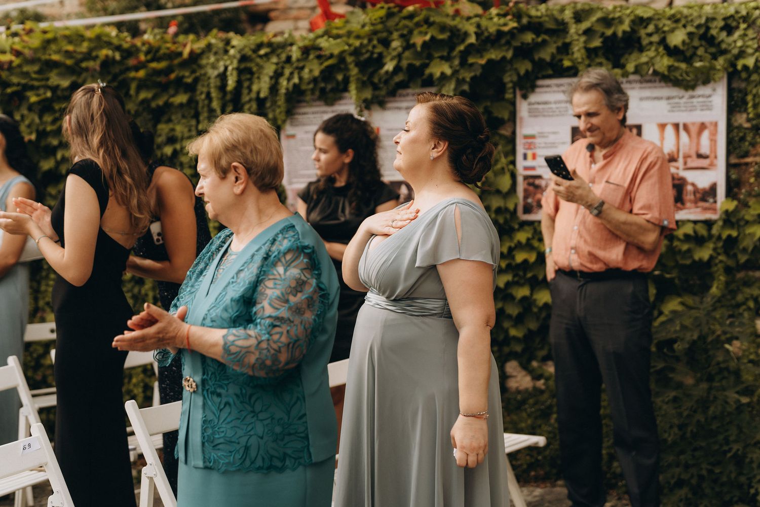 Guests in formal attire gather outdoors at a wedding ceremony, standing near white chairs and a lush green ivy wall.