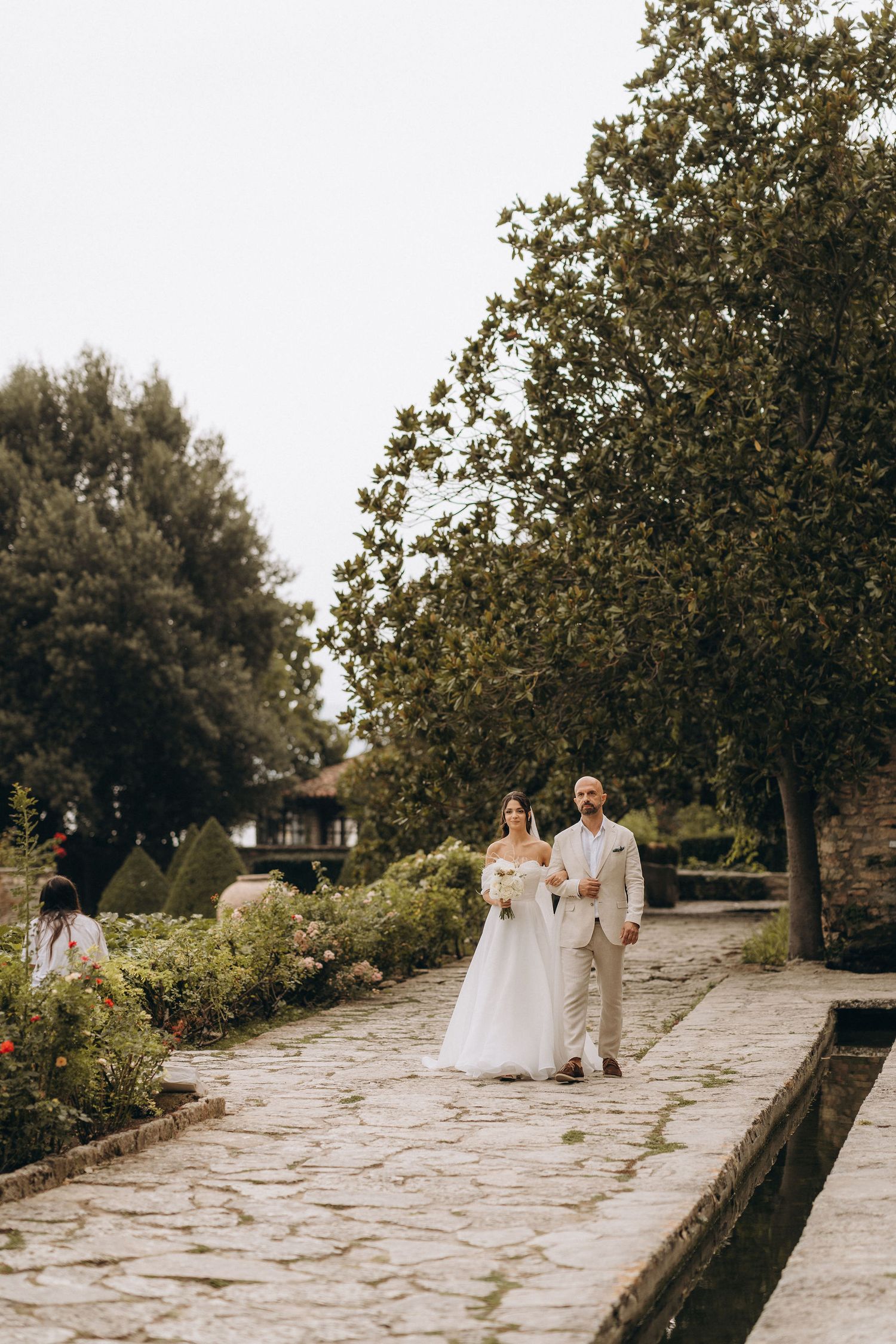 Bride and groom walk hand in hand along a stone garden path surrounded by lush green trees and flowers.