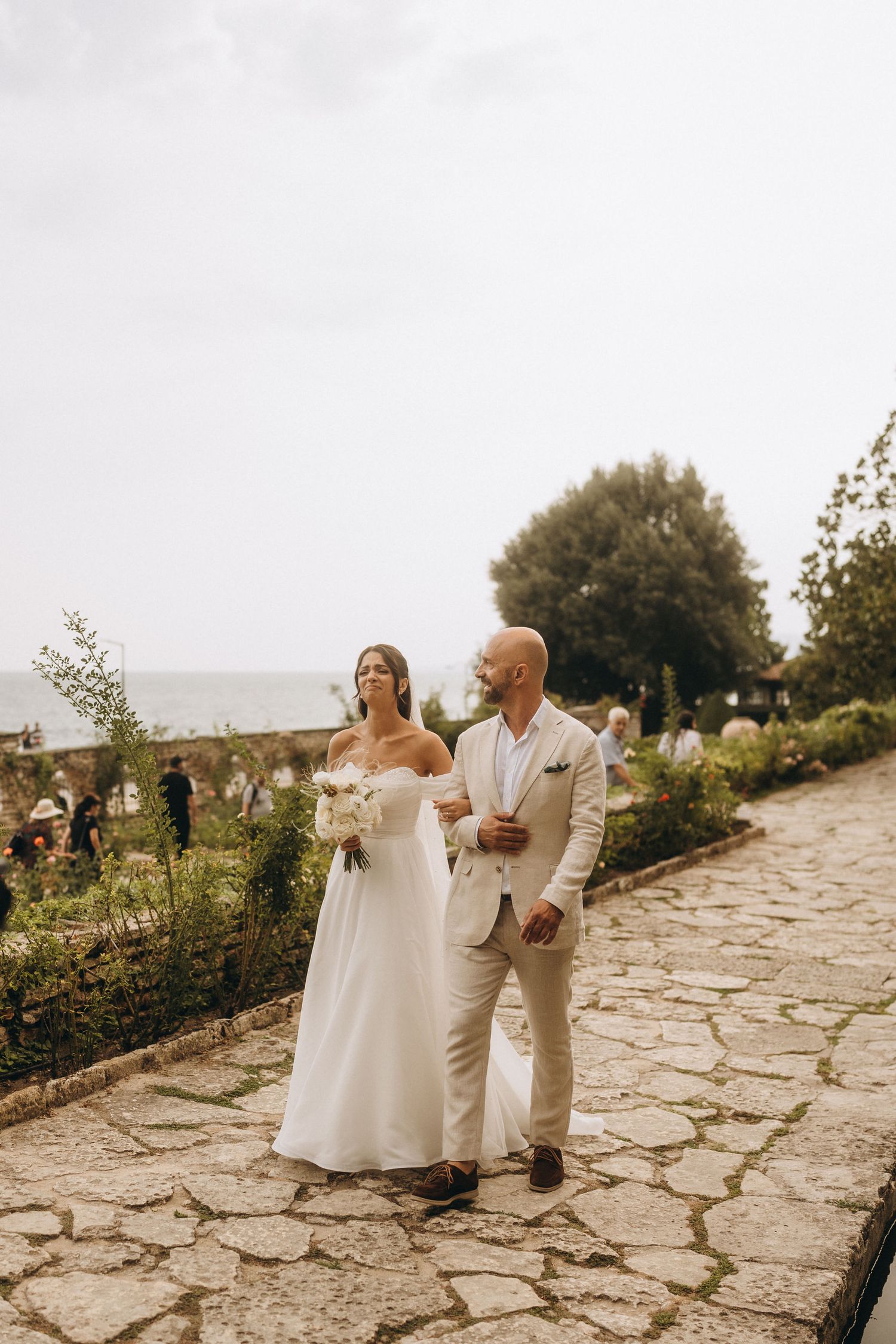Bride and groom walking hand in hand on a stone path in a lush garden with ocean views on their wedding day.