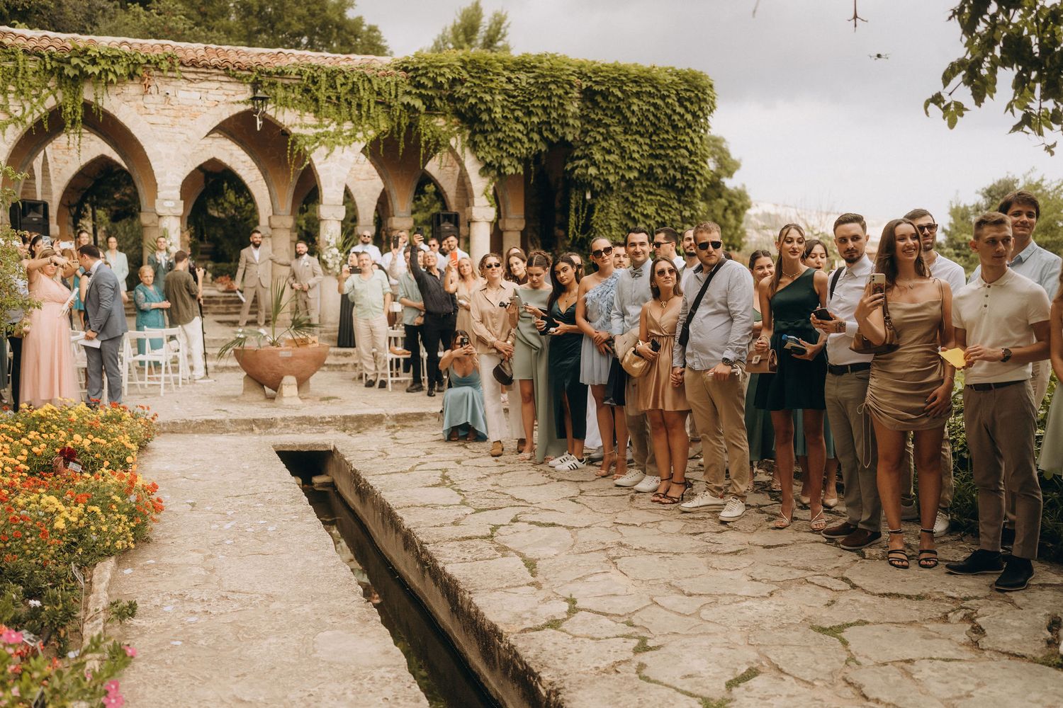 Guests gather outdoors at a rustic stone venue with ivy-covered arches for an elegant garden wedding celebration.