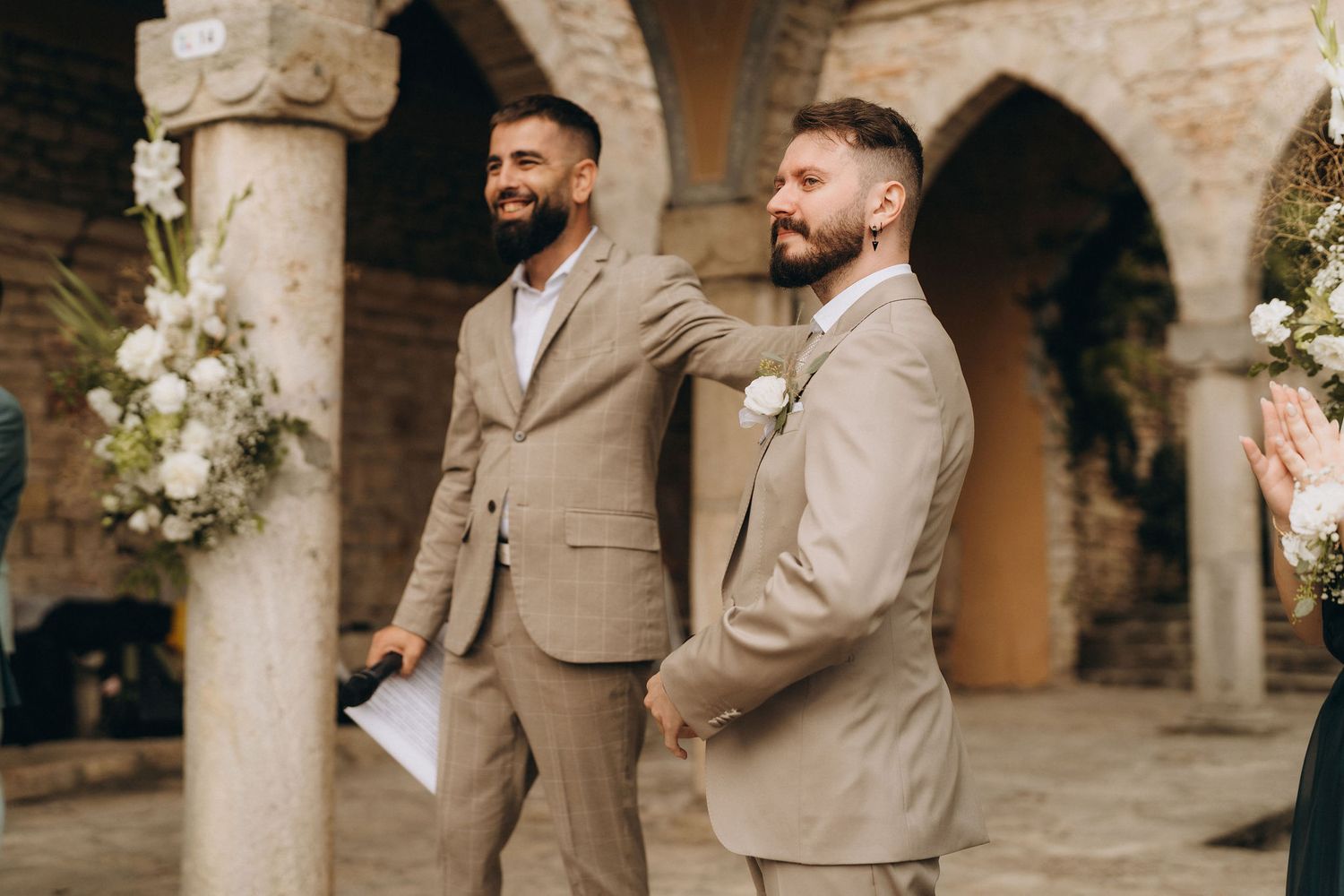 Two grooms in matching tan suits smile during an outdoor wedding ceremony with stone columns and floral decor.