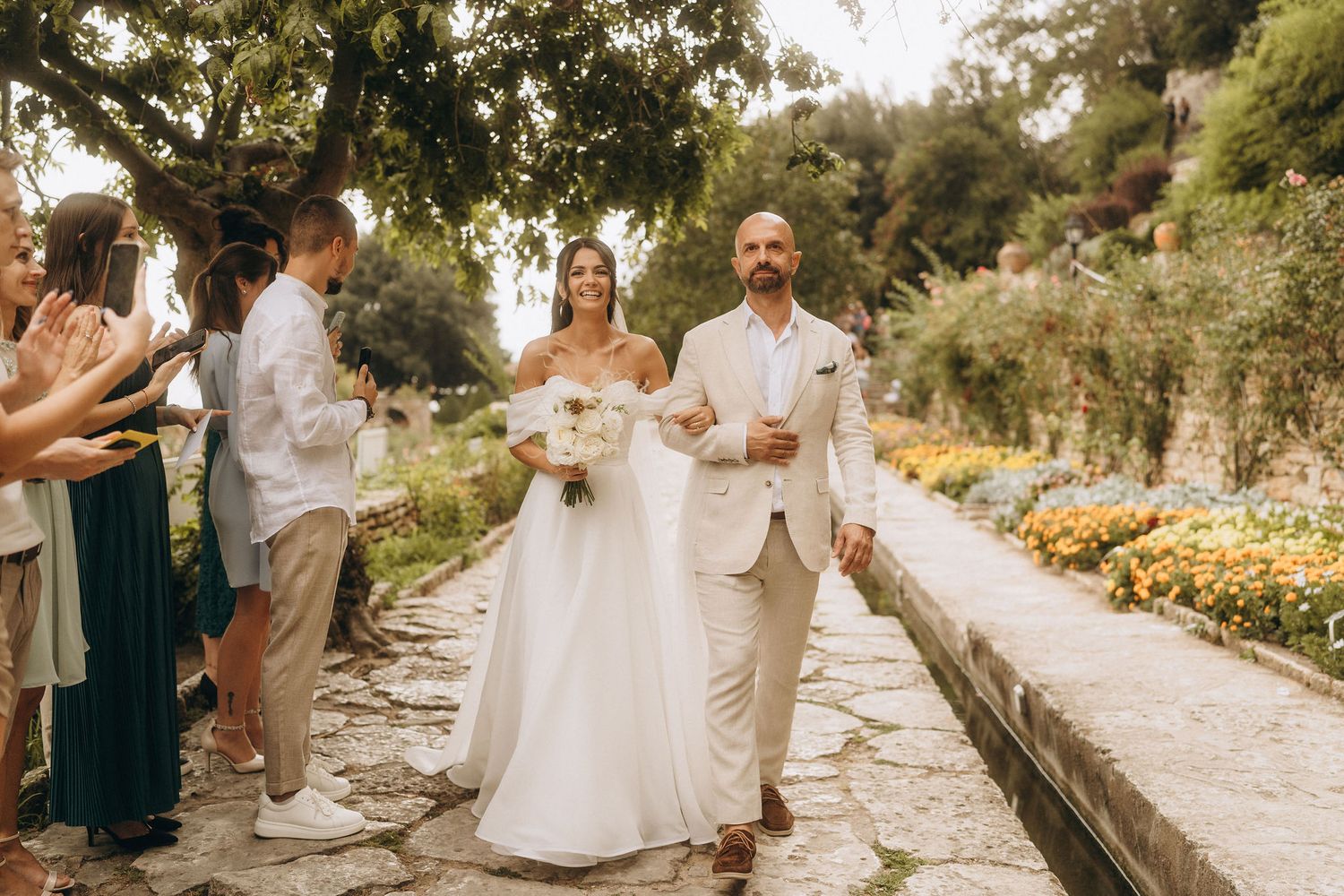 Bride and groom walk down outdoor garden aisle surrounded by cheering guests at a sunny wedding ceremony.