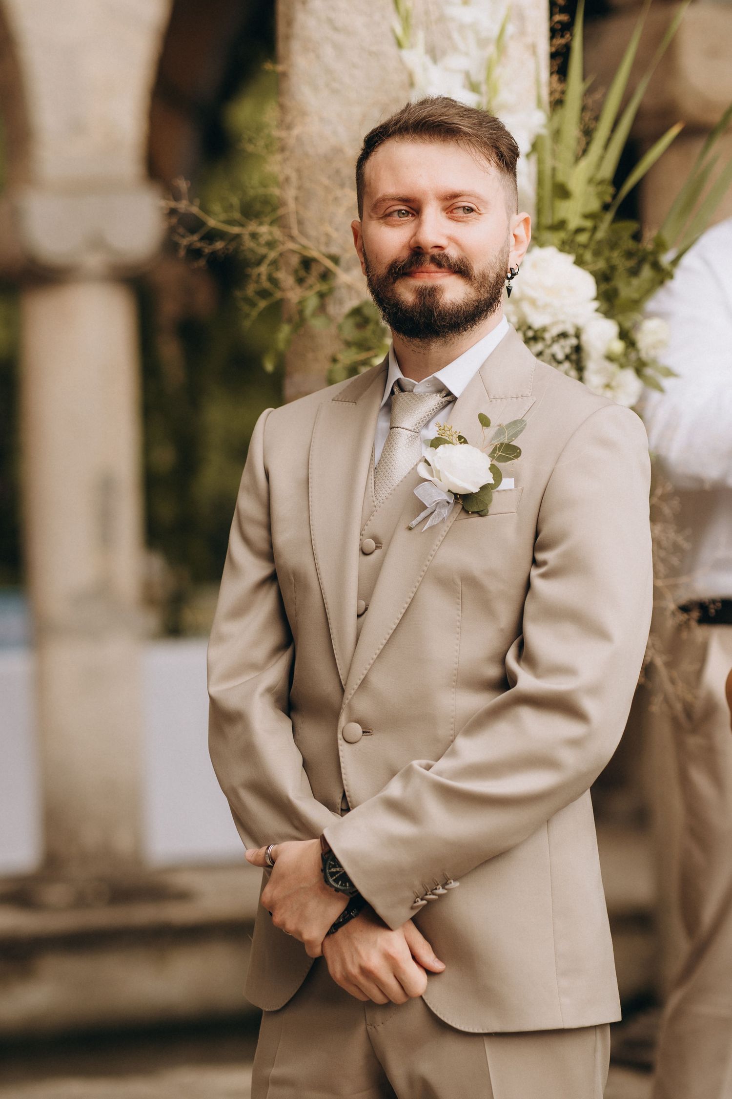 Bearded groom in beige suit with white rose boutonniere smiling at outdoor wedding ceremony.