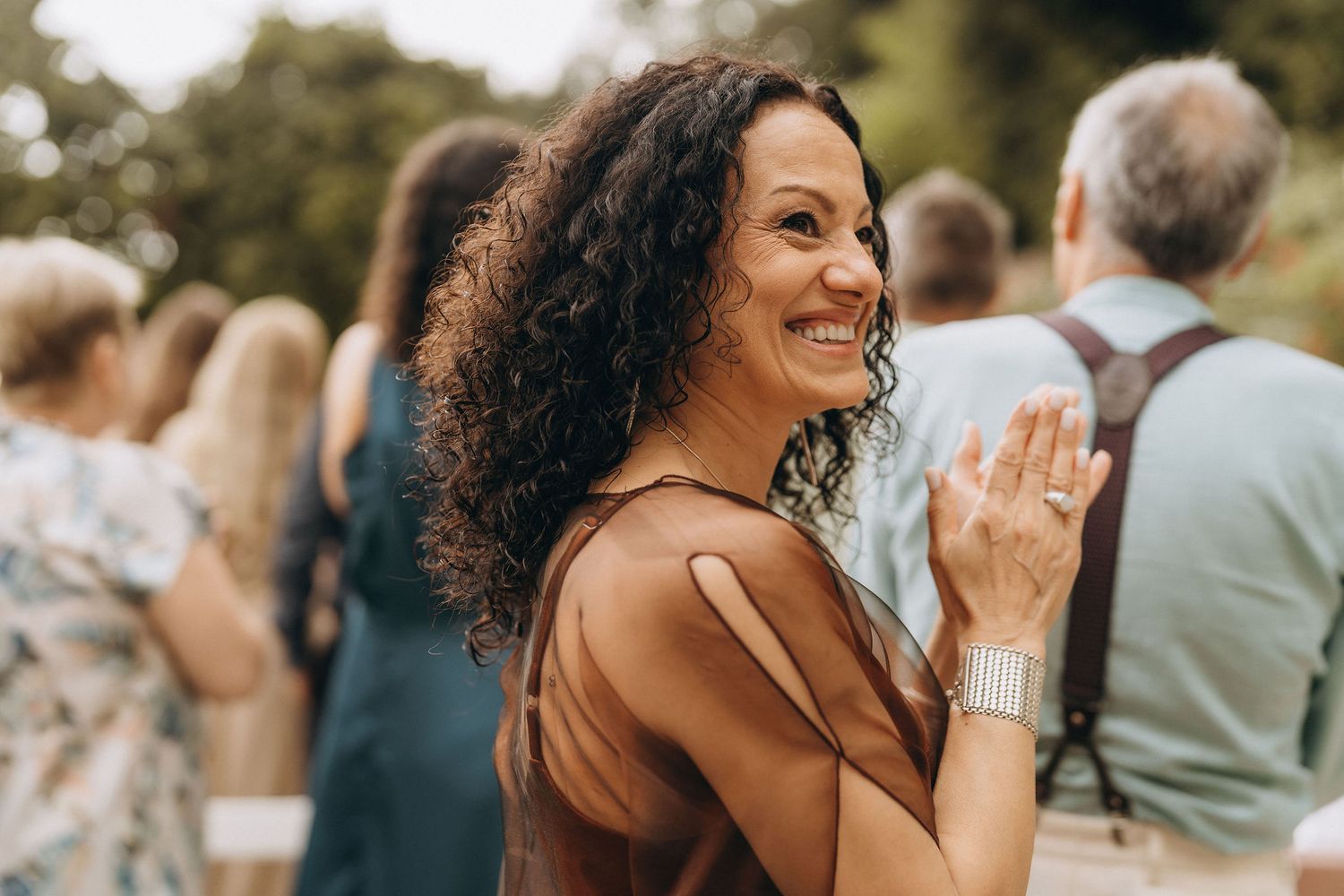 A smiling woman with curly hair in a brown dress turns back at an outdoor gathering event.