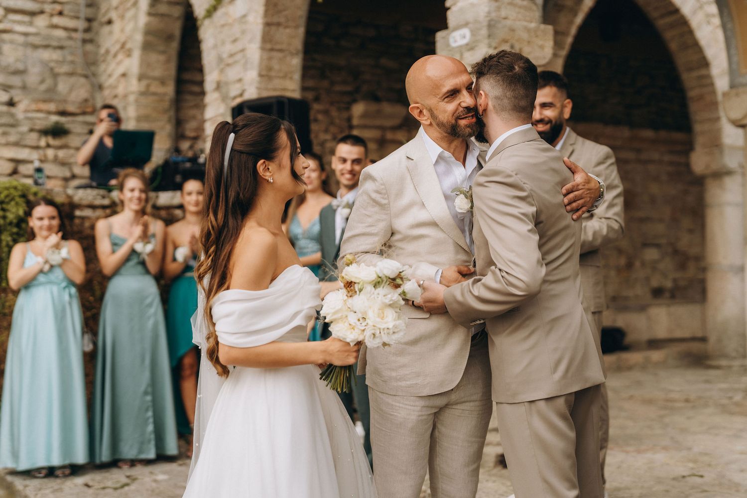 Groom embracing best man at outdoor wedding ceremony while bride holds white bouquet, bridesmaids in teal dresses behind.