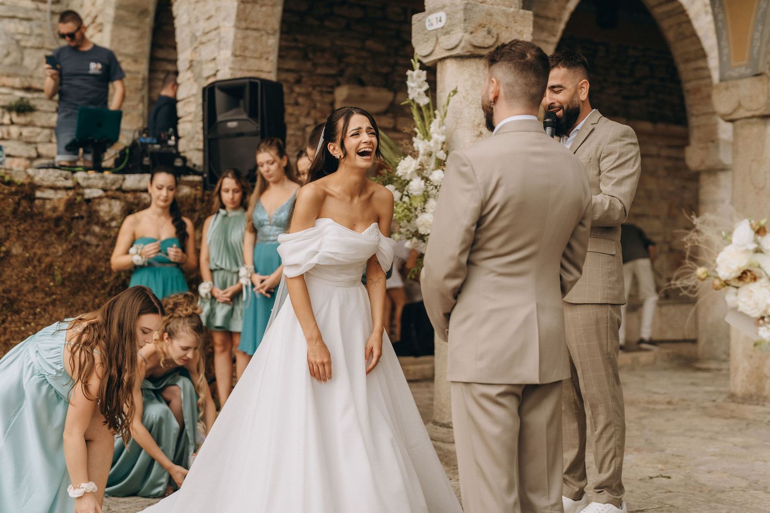 Bride laughing joyfully with groom at outdoor wedding ceremony while bridesmaids in teal dresses adjust her white ballgown.