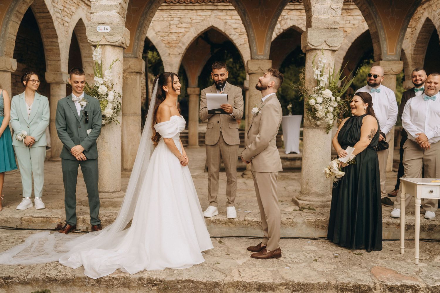 Elegant outdoor wedding ceremony under stone arches with bride in white gown and groom in tan suit.