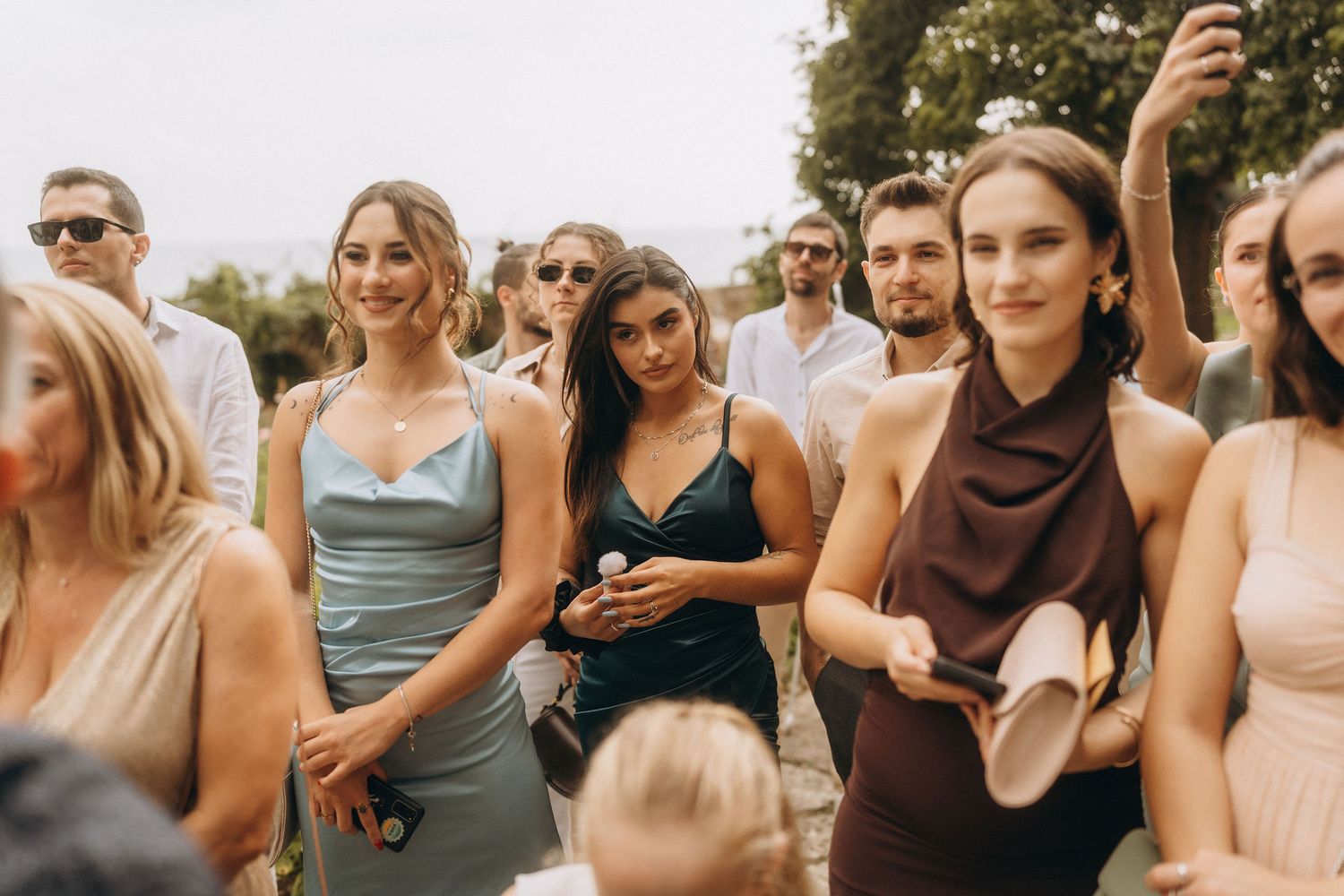 Guests in elegant attire smile and celebrate at an outdoor wedding reception on a sunny day.