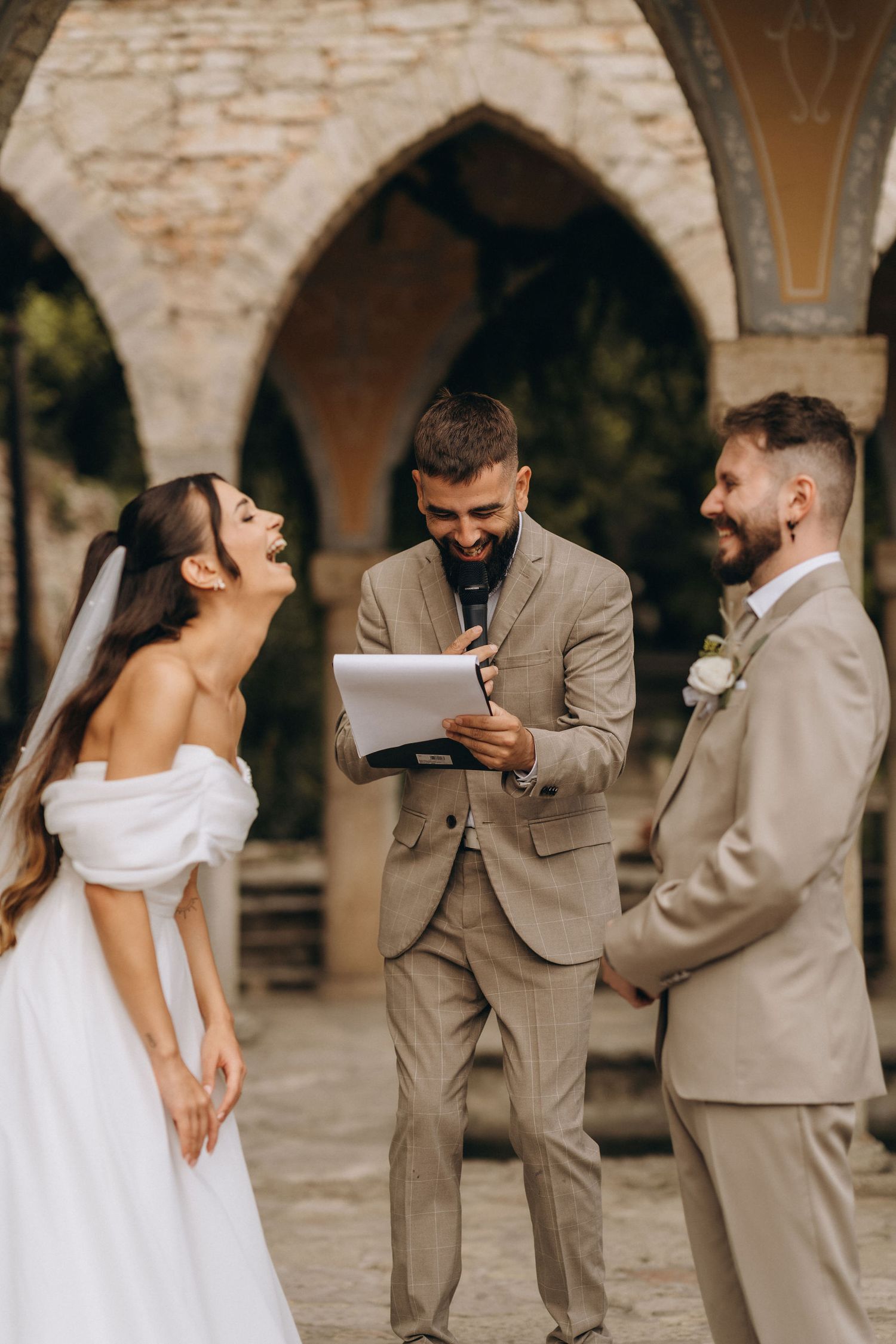 Bride and groom laughing with officiant during outdoor wedding ceremony at historic stone arch venue.