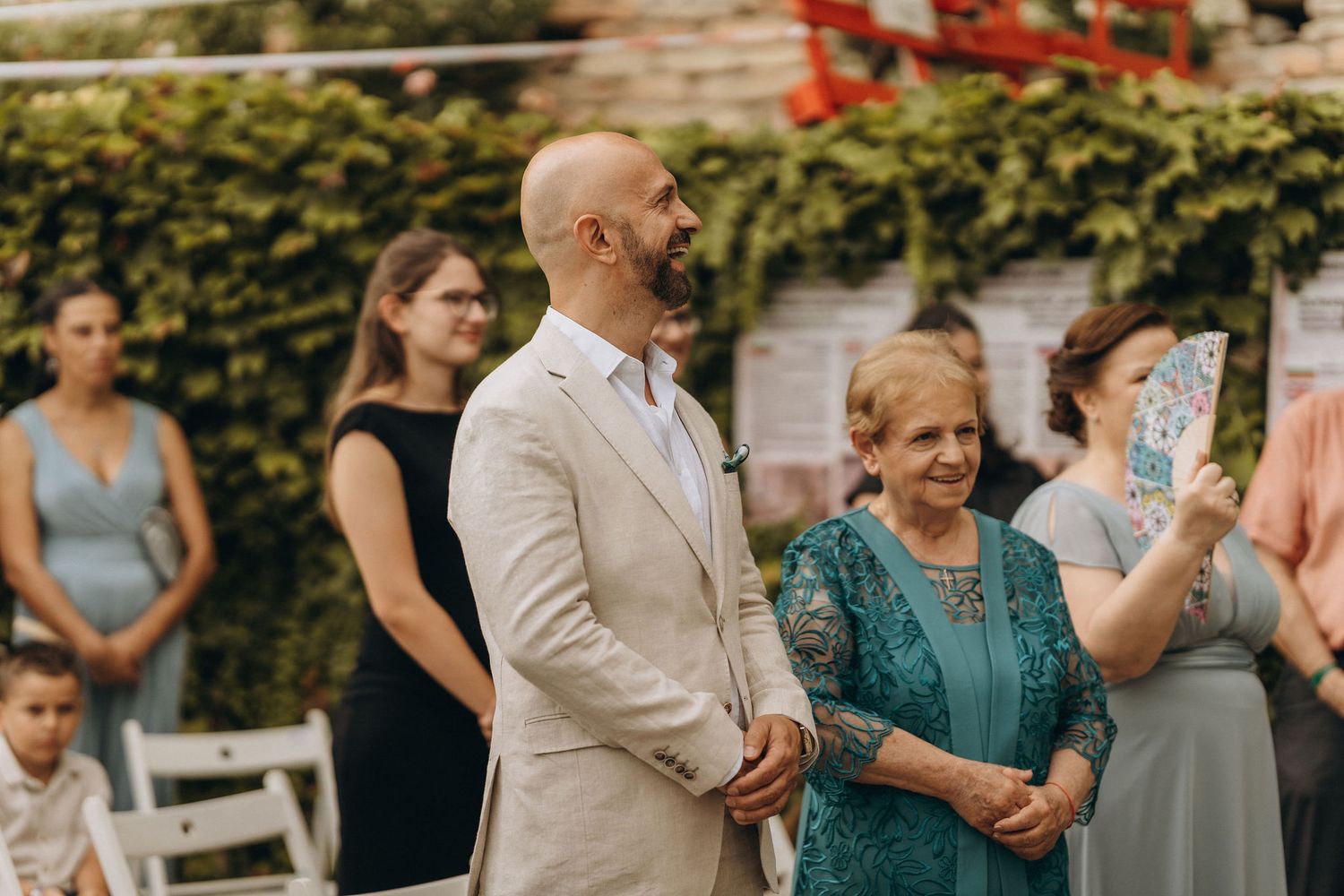 Bald groom in beige suit smiling at wedding ceremony beside elderly woman in teal dress outdoors.