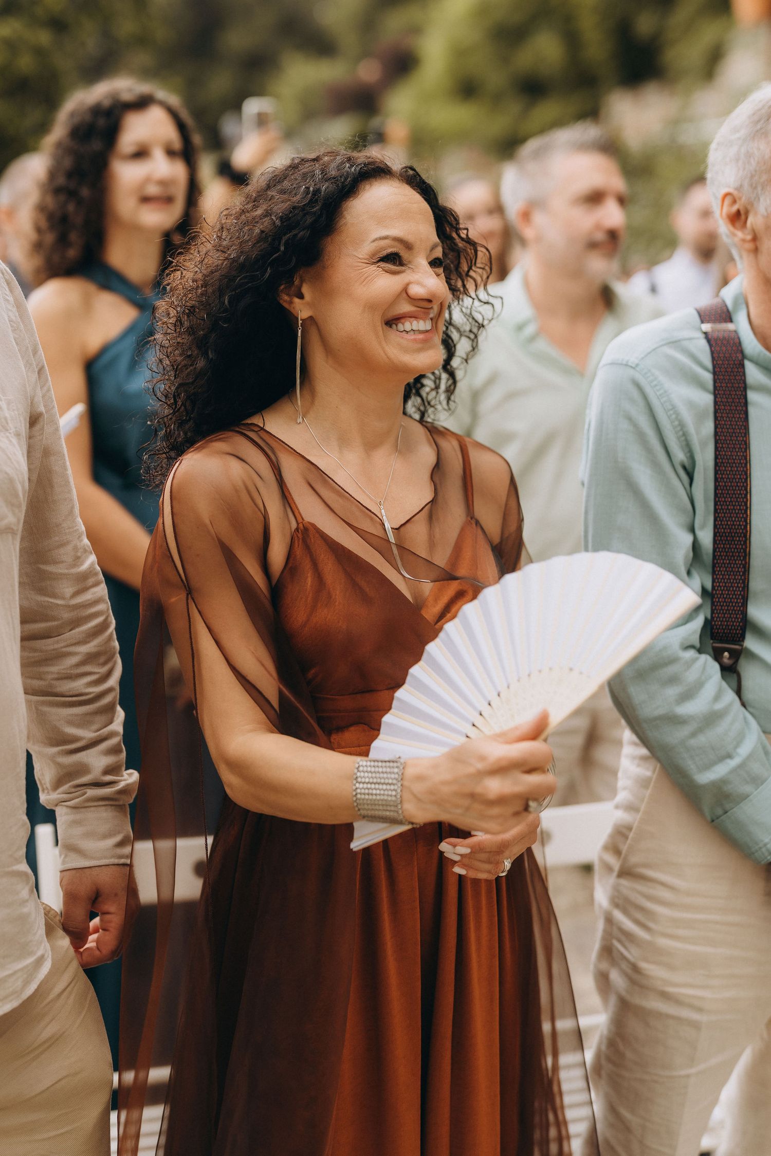 Smiling woman in brown dress holding white hand fan at outdoor social gathering.
