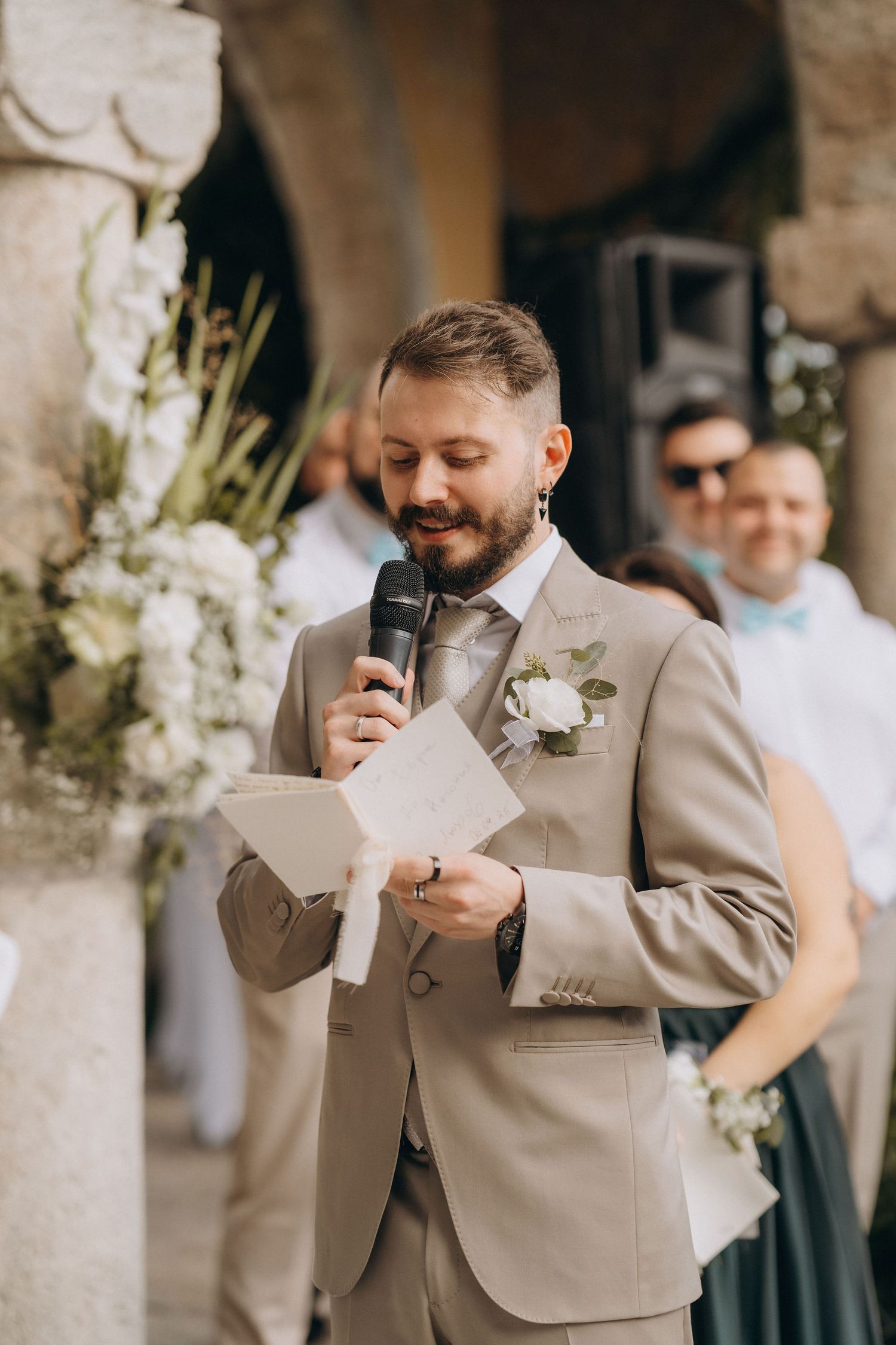 Bearded groom in beige suit reads vows into microphone with white boutonniere at outdoor wedding ceremony.