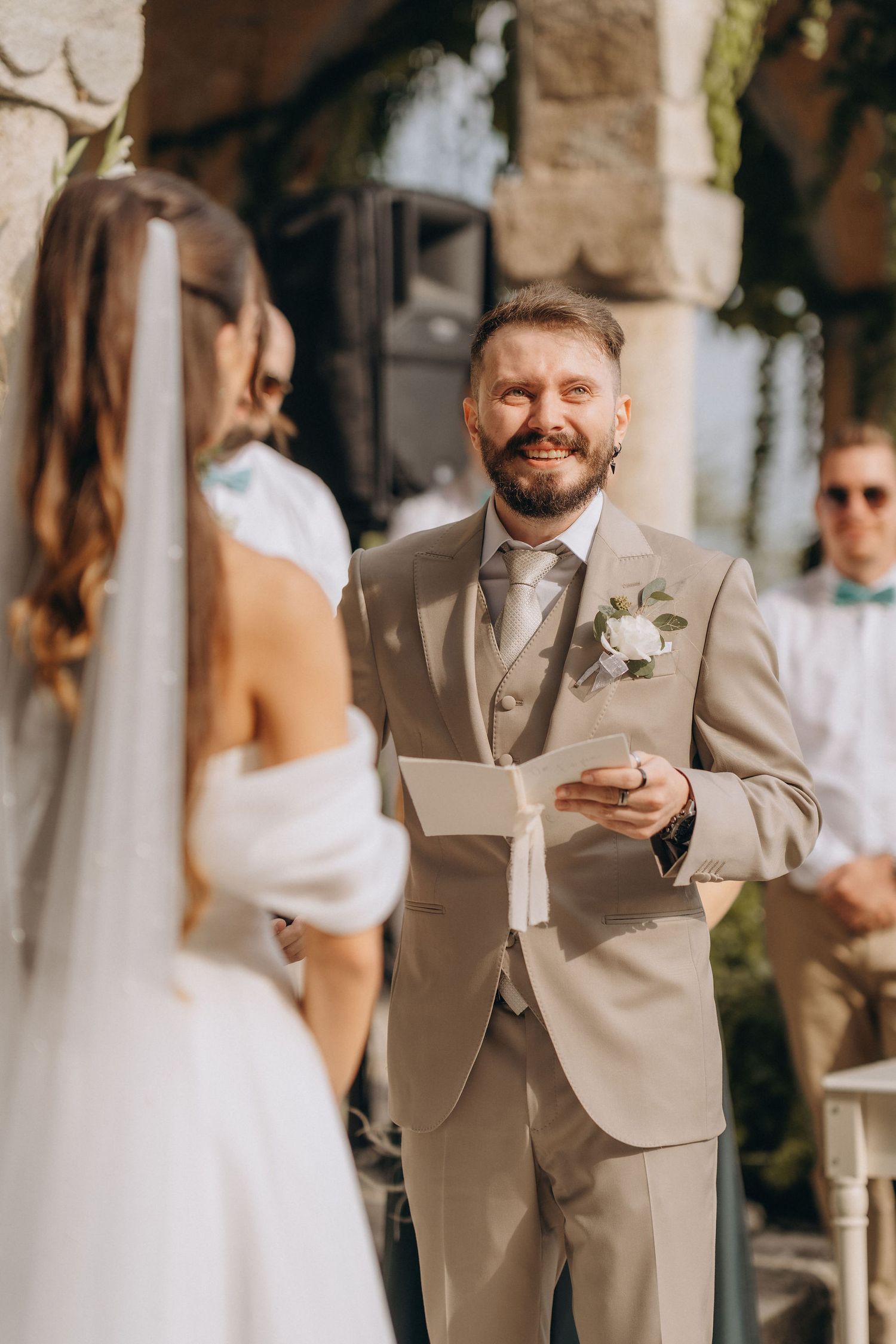 Smiling groom in beige suit reads wedding vows to bride in white dress during outdoor ceremony.