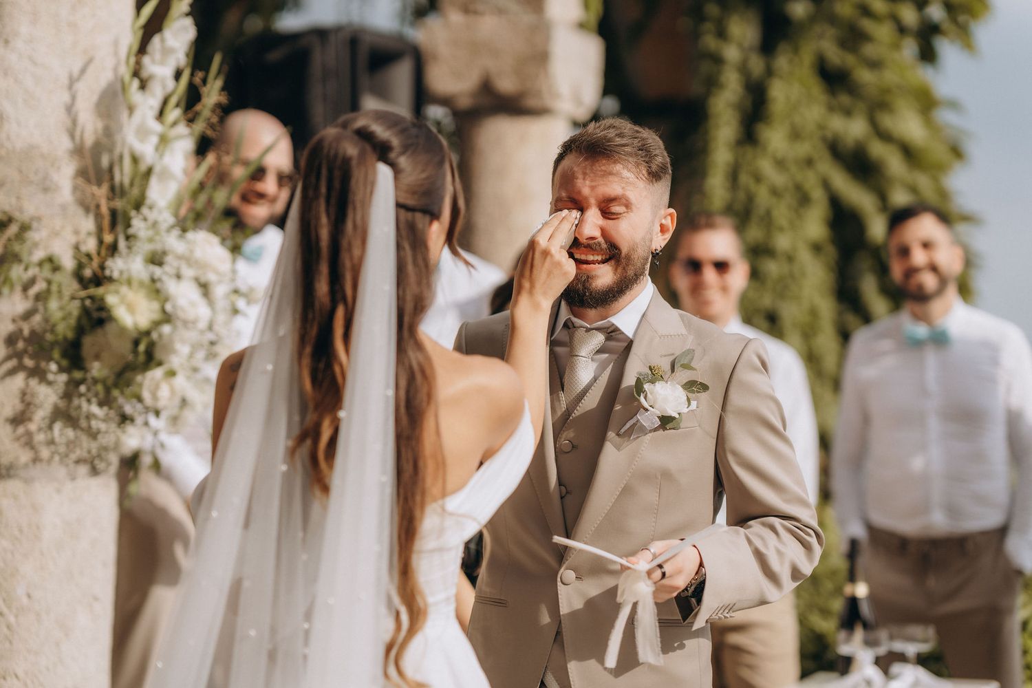 Bride wiping tears from groom's cheek during outdoor wedding ceremony, groom in beige suit with white boutonniere.